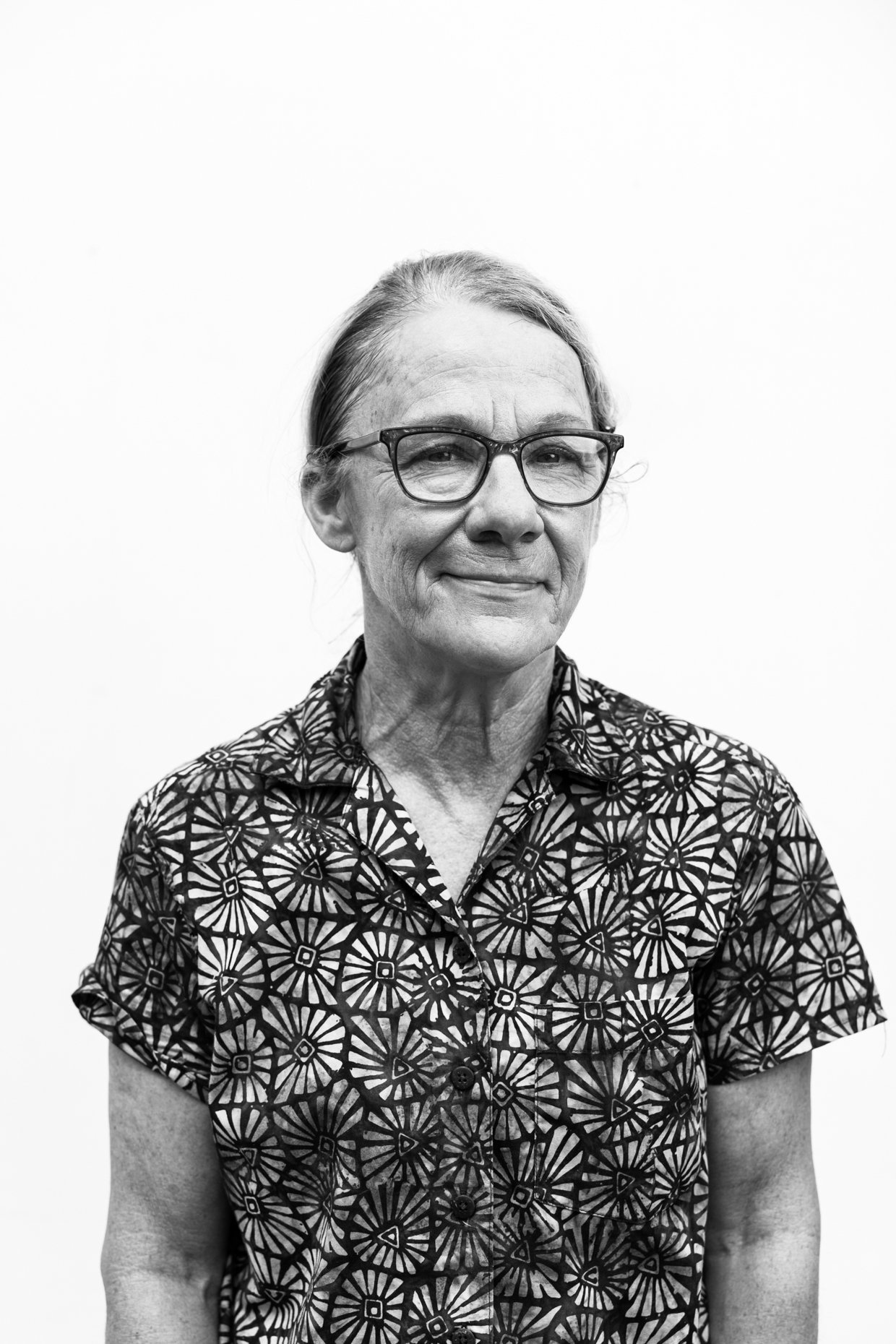 Black and white portrait of an older woman with glasses, wearing a patterned short-sleeve shirt, smiling subtly, facing the camera against a plain background.