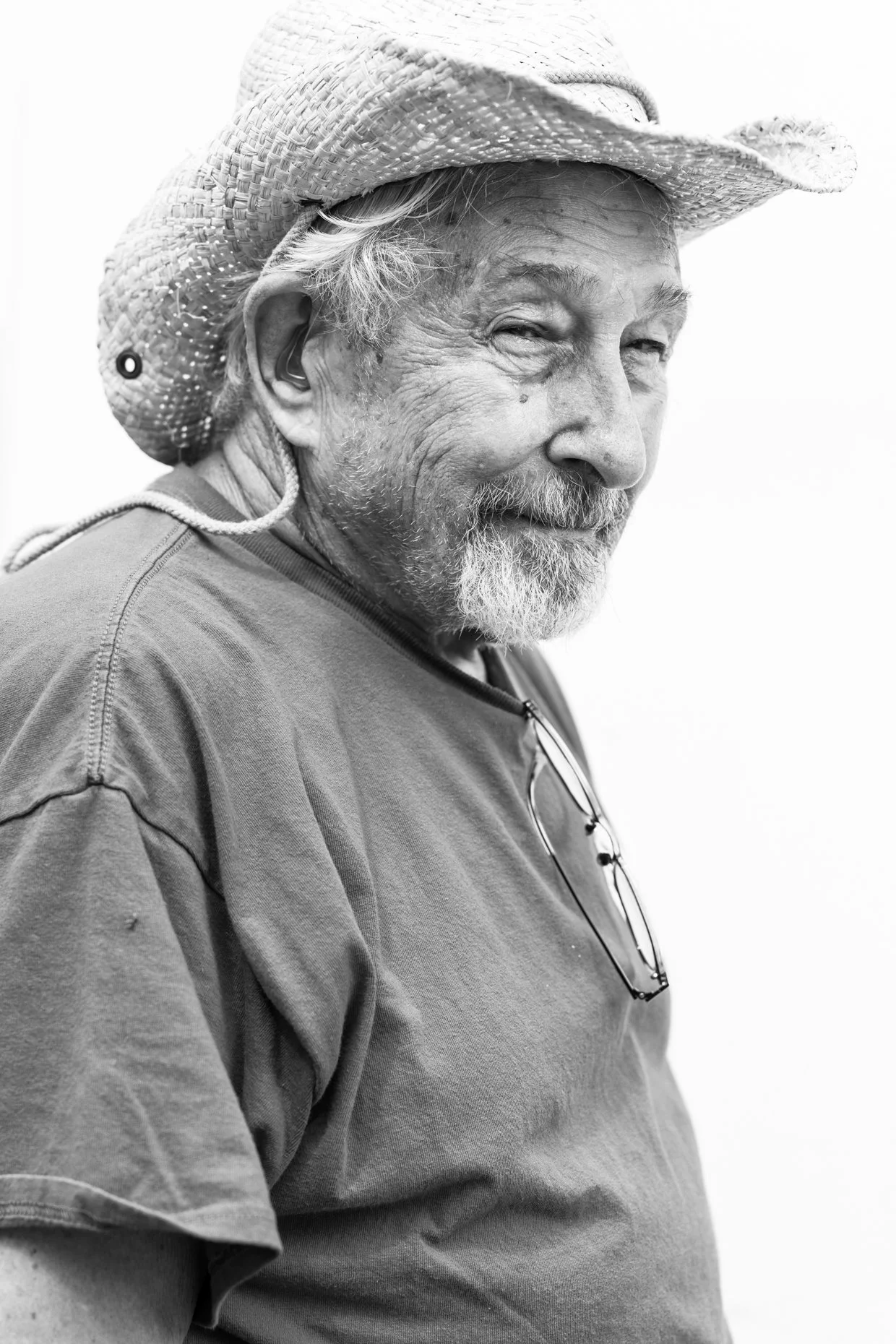 A black and white photograph of an older man wearing a wide-brimmed straw hat and casual t-shirt, with a pair of glasses hanging from his shirt collar. He has a beard, mustache, and a gentle smile.