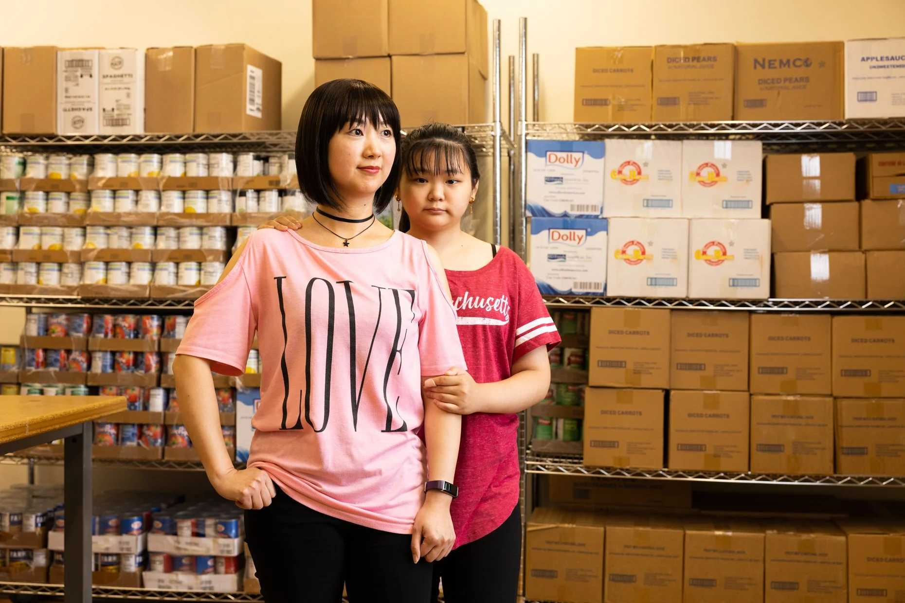 Two young women standing close together in a storage room with shelves of canned goods and boxes. One woman is wearing a pink shirt with the word 'LOVE' and a star choker. The other woman is in a red shirt with white lettering. They are posing for a 