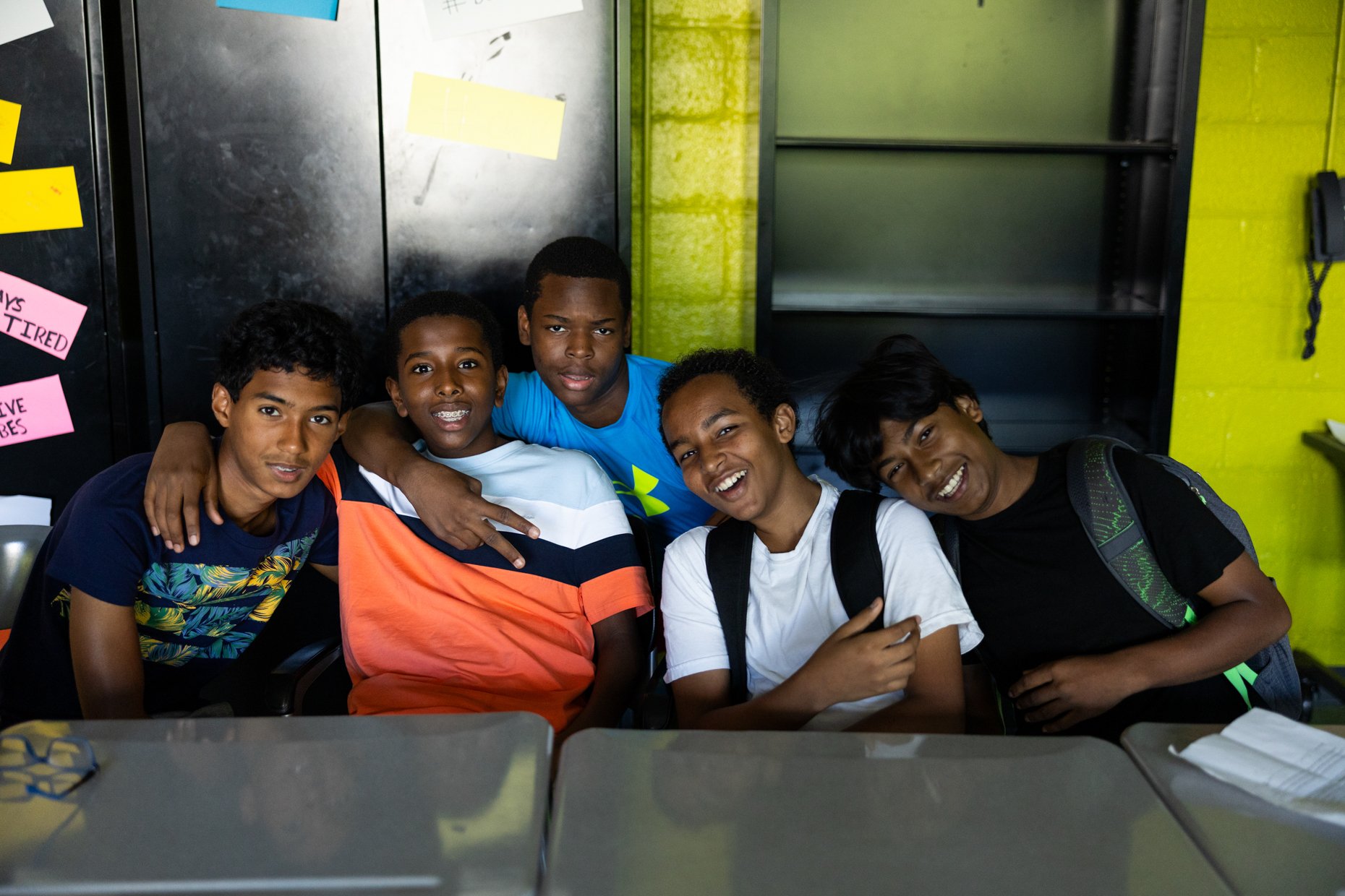 A group of five young boys smiling and posing together in a classroom setting, with some making gestures. They are seated at a table with books and other classroom items visible.