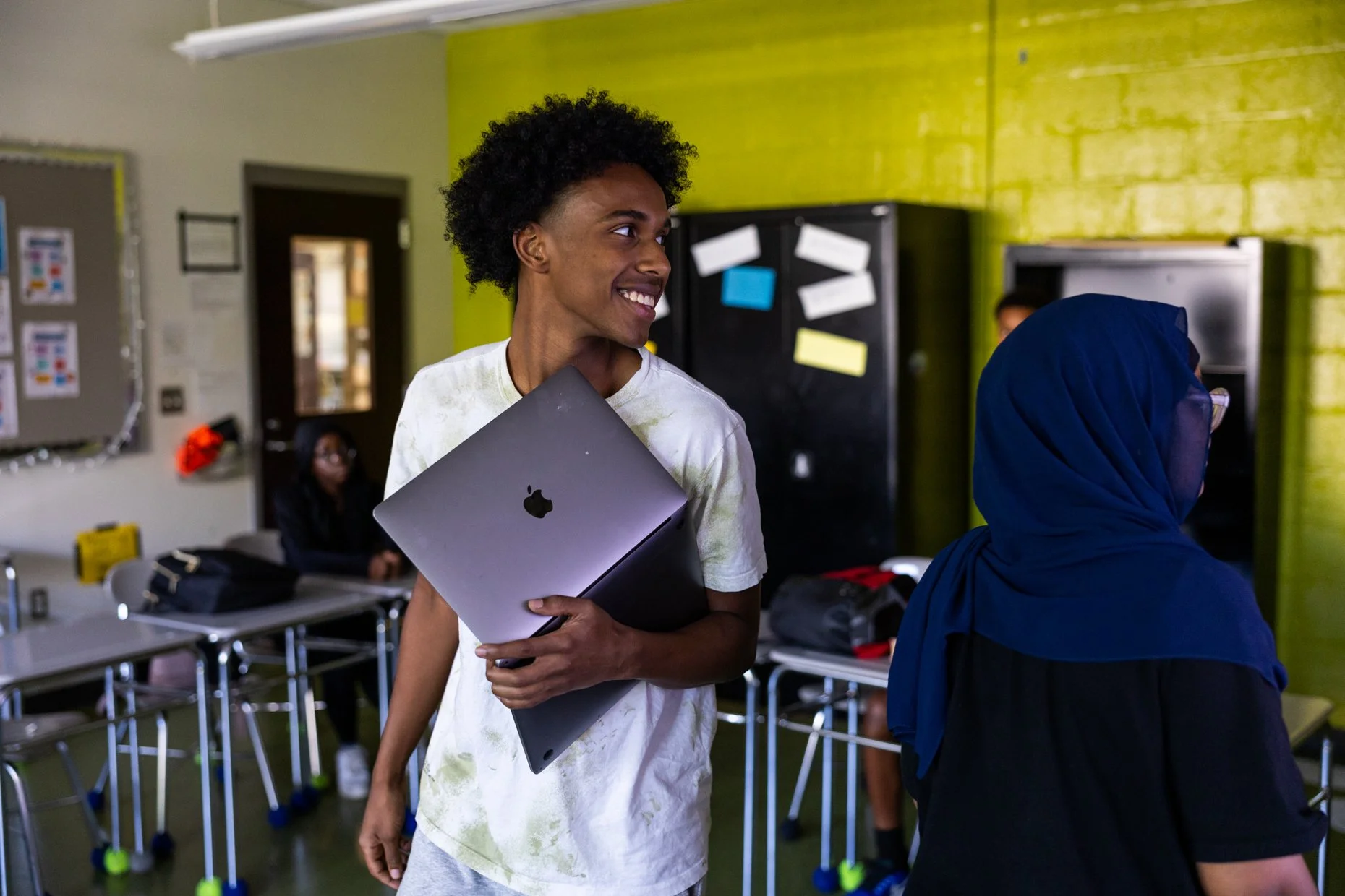 A young man with curly hair smiling and holding a closed laptop in his arms, standing in a classroom with other students and tables.