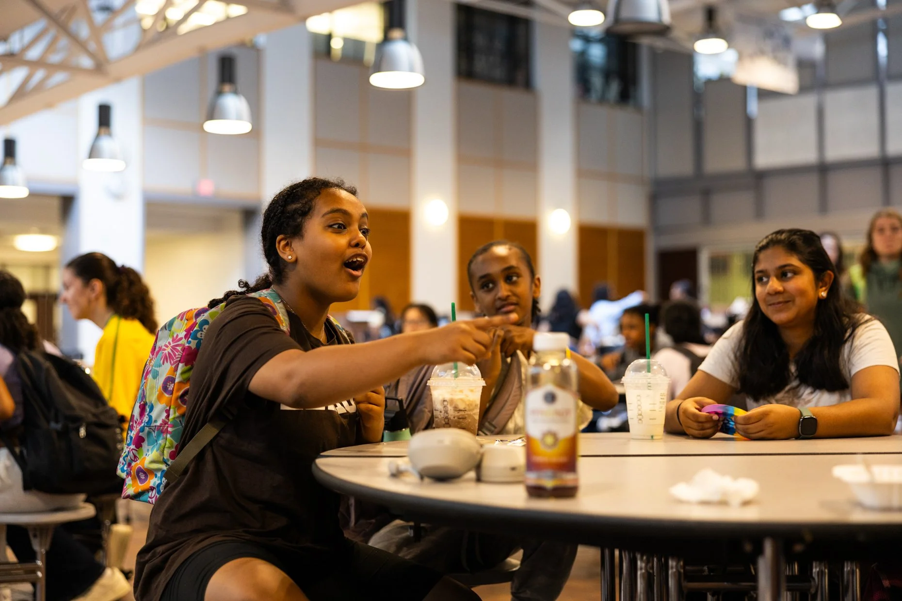 Three young women sitting at a table in a busy modern indoor café or food court, engaging in conversation with drinks and snacks in front of them.