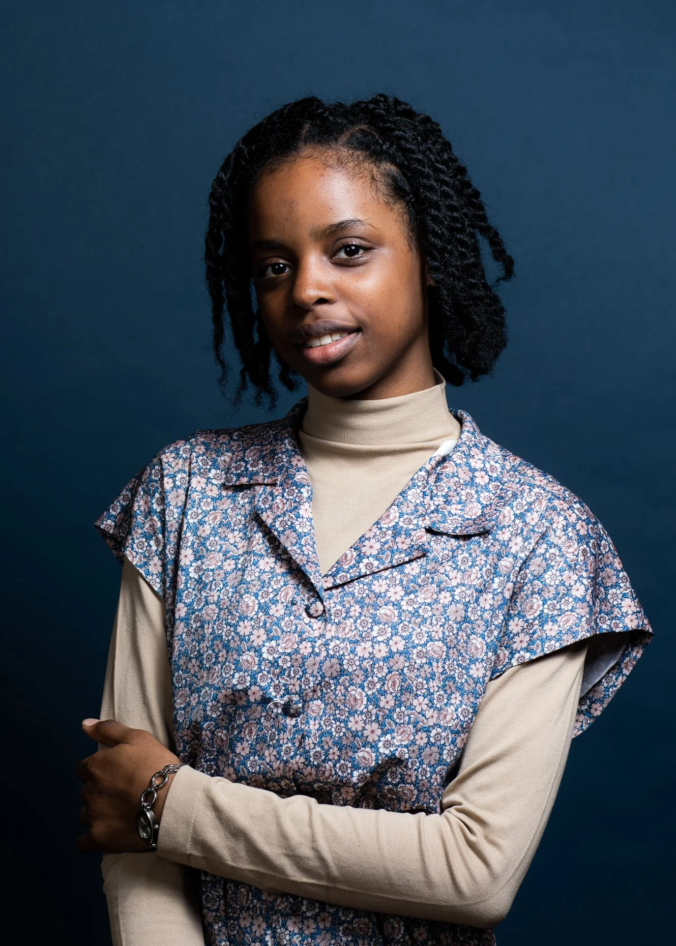 A young woman with styled black curly hair, wearing a beige turtleneck under a floral blouse, standing against a dark blue background with her arms crossed, looking at the camera with a slight smile.