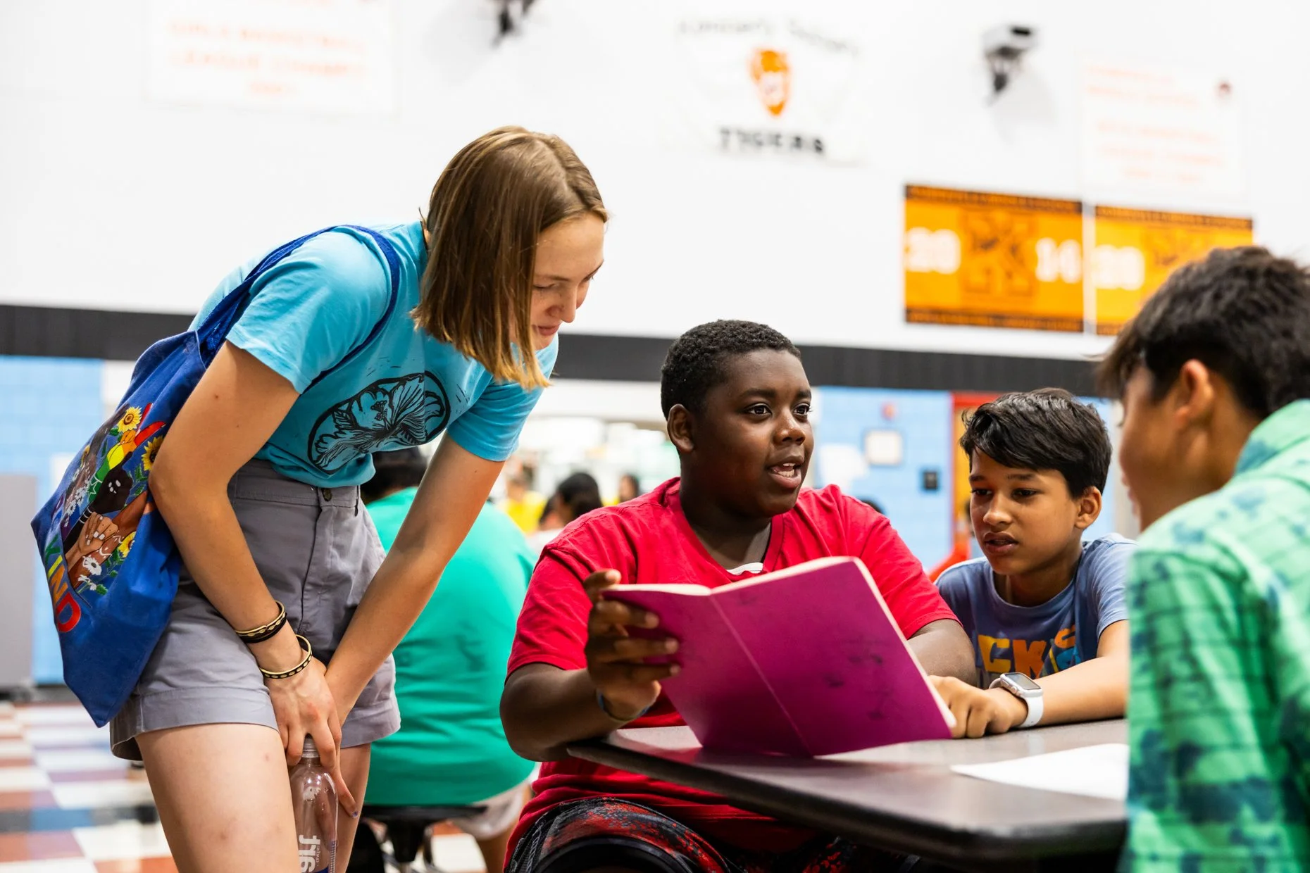A group of four diverse teenagers gathered around a table in a school hallway, looking at a pink notebook with curiosity and interest. One girl with light skin and brown hair, wearing a blue t-shirt and carrying a blue backpack, is leaning over to lo