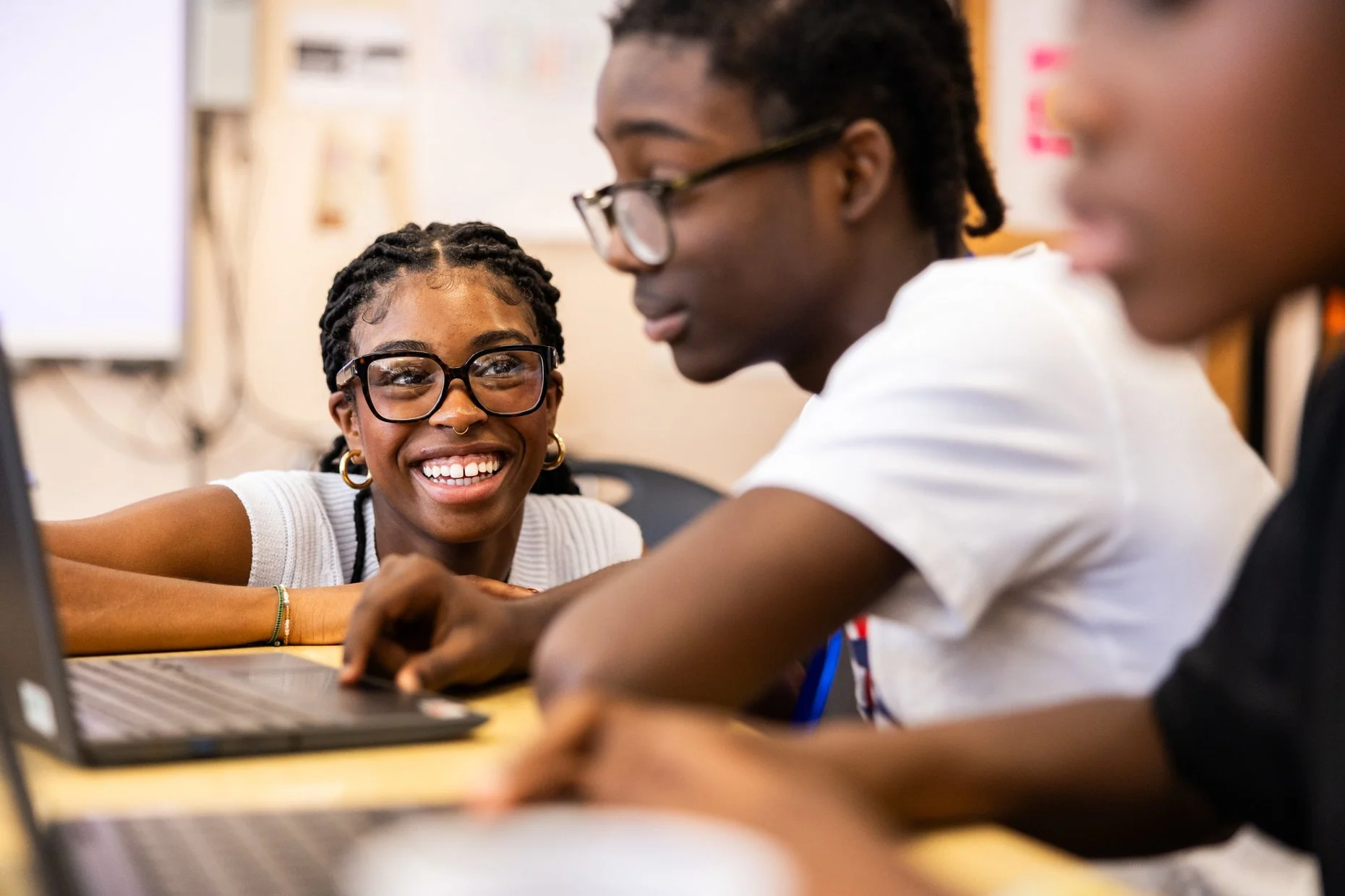Three young people sitting at a table in a classroom, smiling and engaging with each other, with a laptop in front of them.
