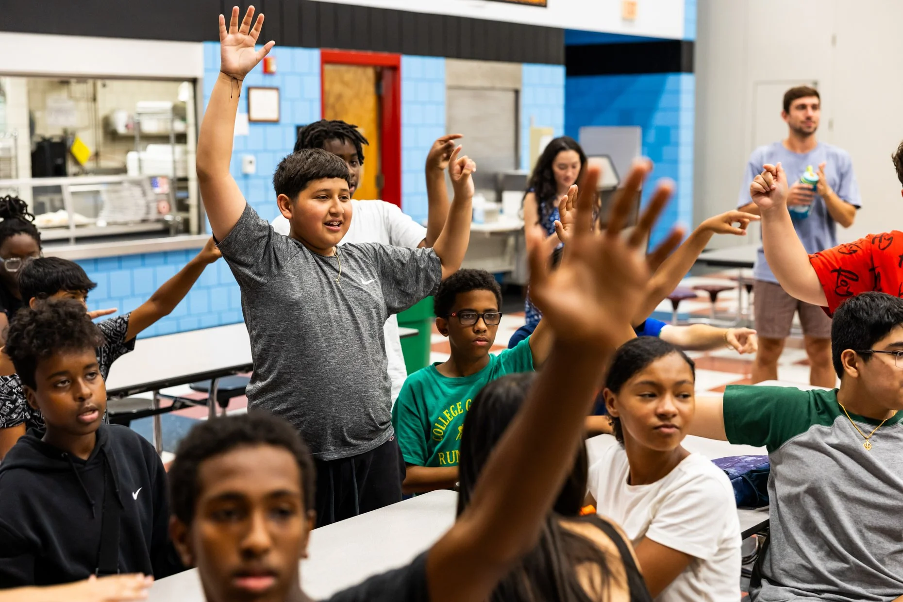 A group of diverse children sitting at tables, participating in a classroom activity with some raising their hands, and an adult standing in the background with a water bottle, in a colorful school cafeteria or classroom.