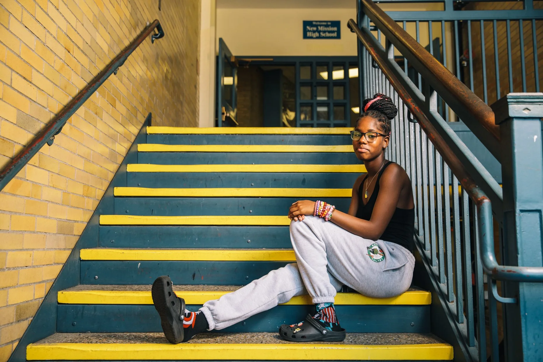 A young girl with glasses and braids, wearing a black tank top, gray sweatpants, and colorful bracelets, is sitting on the yellow and blue staircase at New Minerva High School, beside yellow brick wall and blue railings.