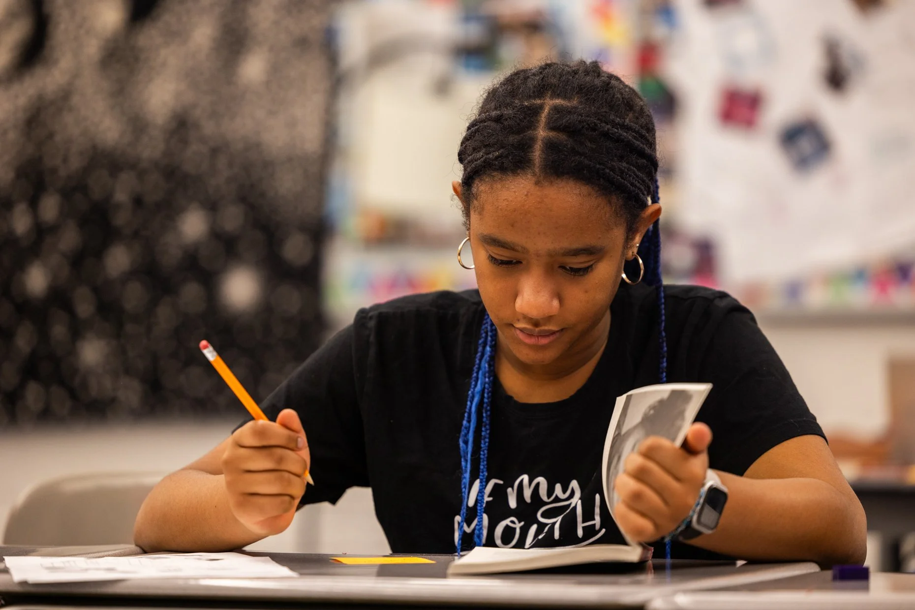 A young woman with braided hair and hoop earrings sitting at a table, holding a photograph in one hand and a pencil in the other, looking at the photo in a classroom.