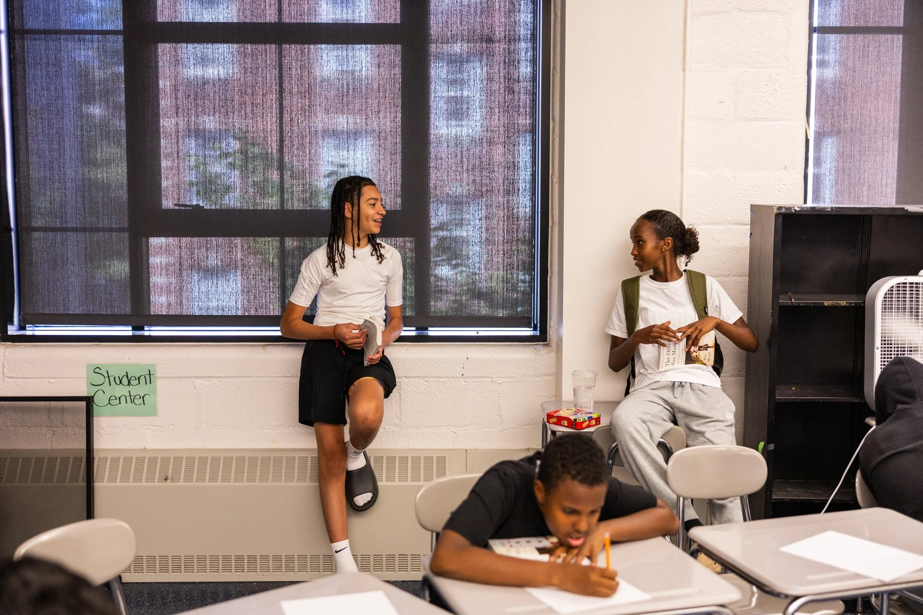 Two young girls standing and sitting in a classroom, engaged in conversation. One girl leans against the wall, holding a hat, while the other sits on a table, holding a book. Several students are seated at desks, working on assignments. A window with