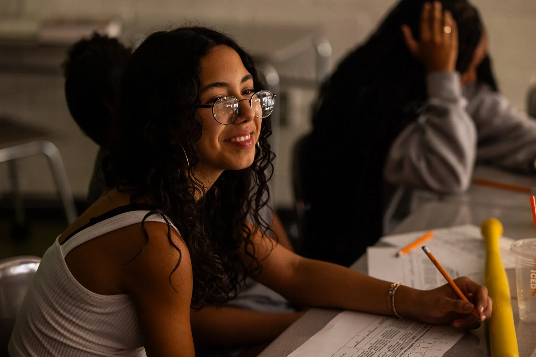A young woman with curly dark hair, glasses, and earrings, smiling and looking to her left. She is sitting at a desk with papers, a pencil, and a yellow container, in a classroom setting with other students.