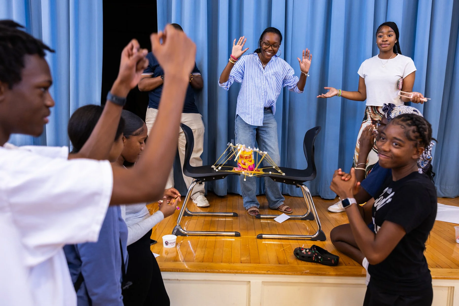 Group of young people gathered around a table with a creative project made of colorful sticks and a bag of snacks. Some are squatting and others are standing, engaging with the project. Blue curtains are in the background.