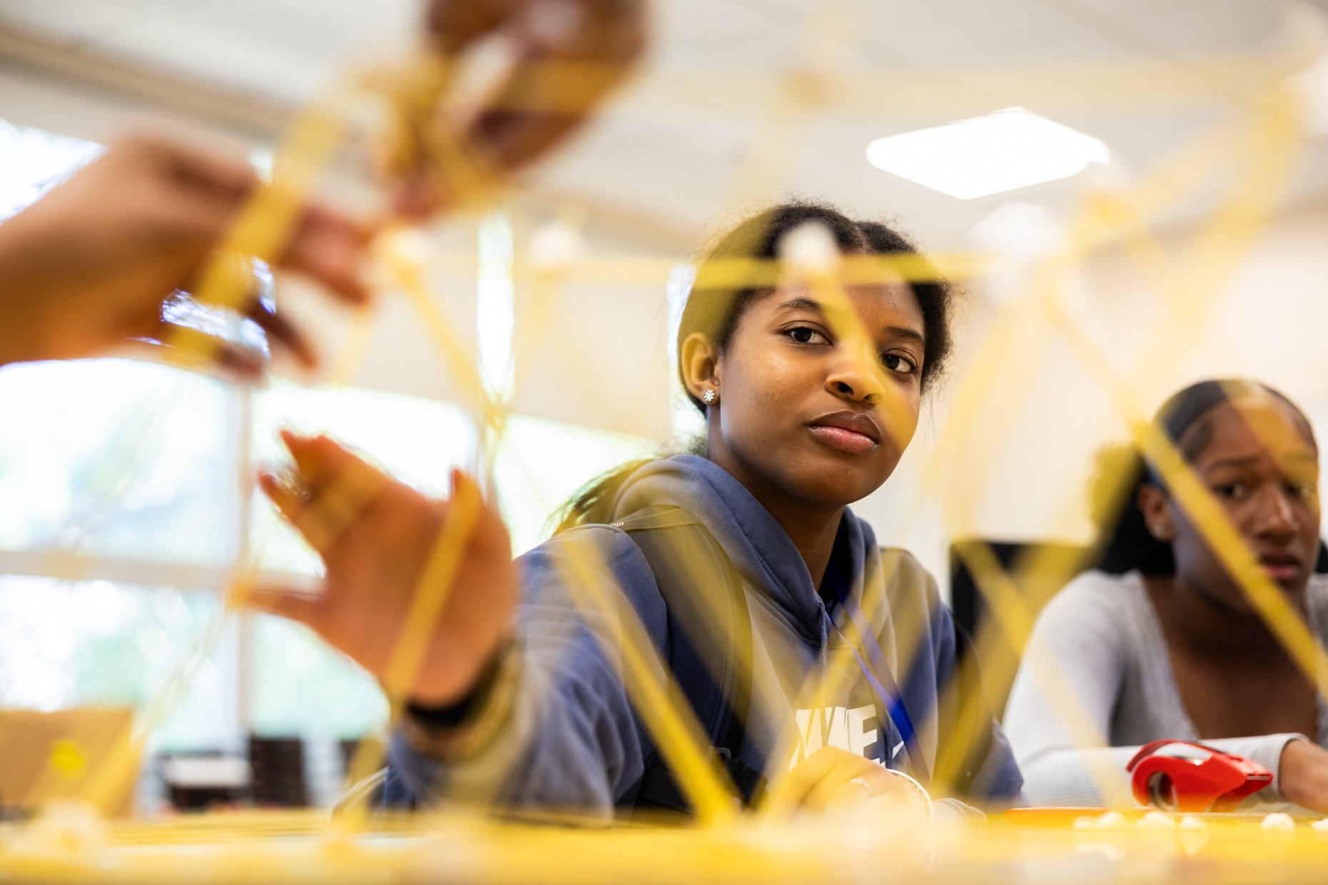 Young woman with dark curly hair and earrings sitting at a table, working on a project with yellow straws and red scissors, in a classroom with bright lighting.