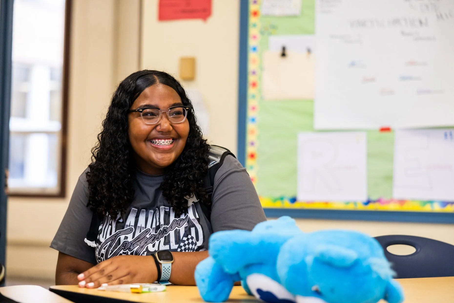 A young woman with curly black hair, glasses, and braces smiling while sitting at a table in a classroom with a colorful bulletin board in the background.