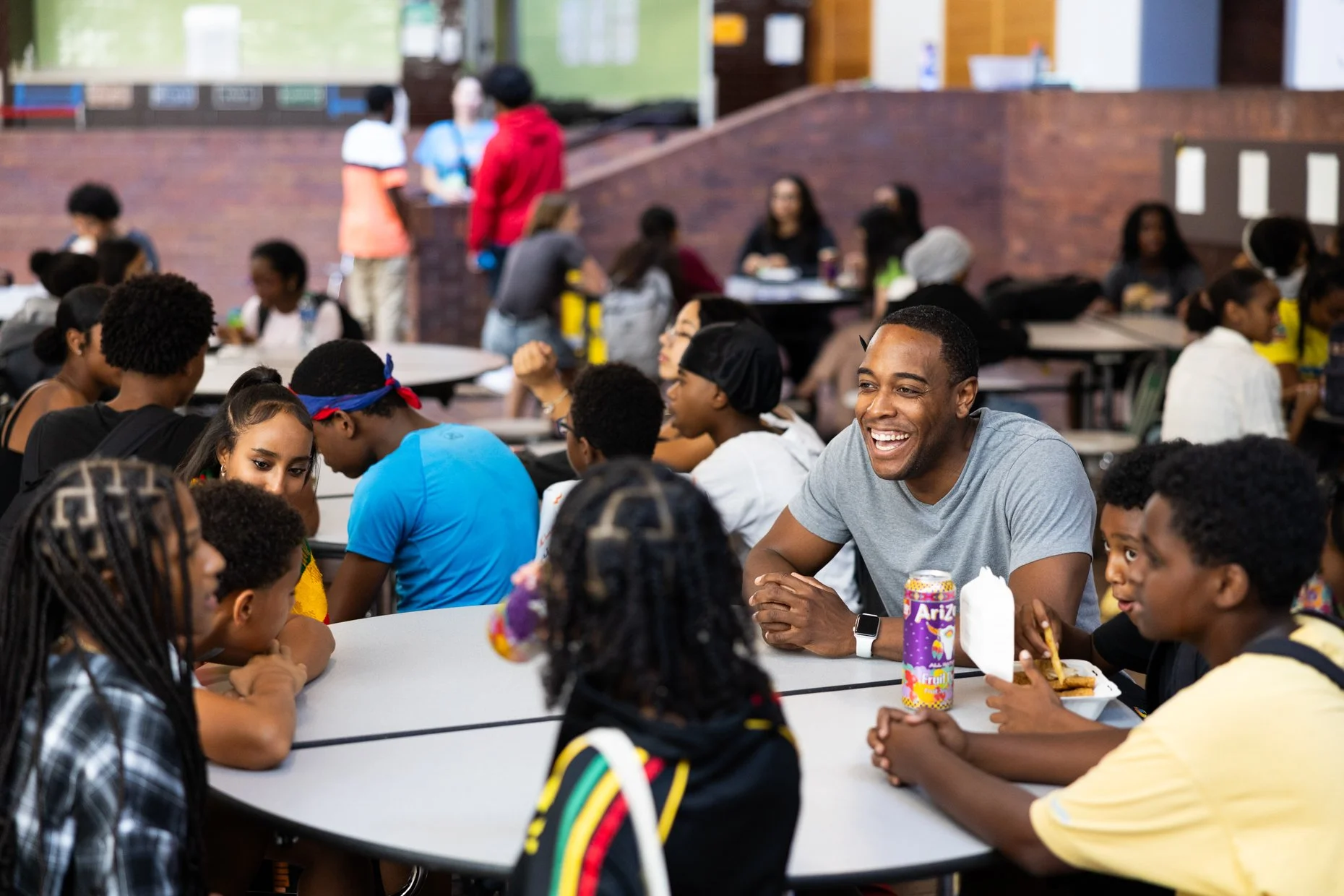 Group of diverse children and teenagers sitting at tables in a school cafeteria, with an adult smiling in the background.