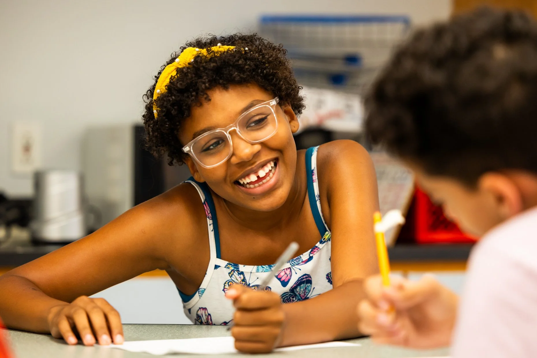 A young girl with short curly hair, wearing glasses, a yellow headband, and a sleeveless top with a colorful butterfly pattern, laughing and interacting with someone in the classroom.