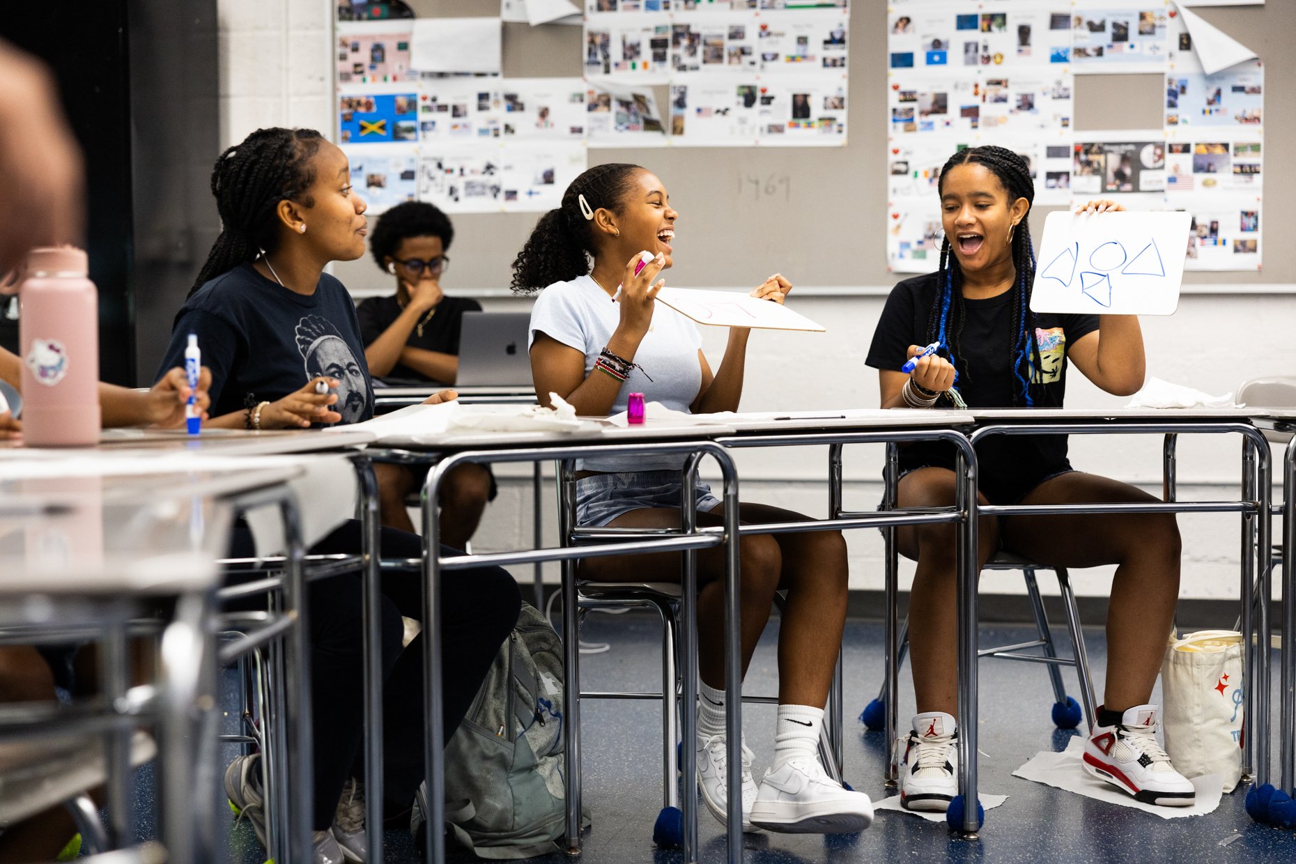 Group of four young women sitting at desks in a classroom, laughing and holding up whiteboards with drawings, with more students and classroom decorations in the background.