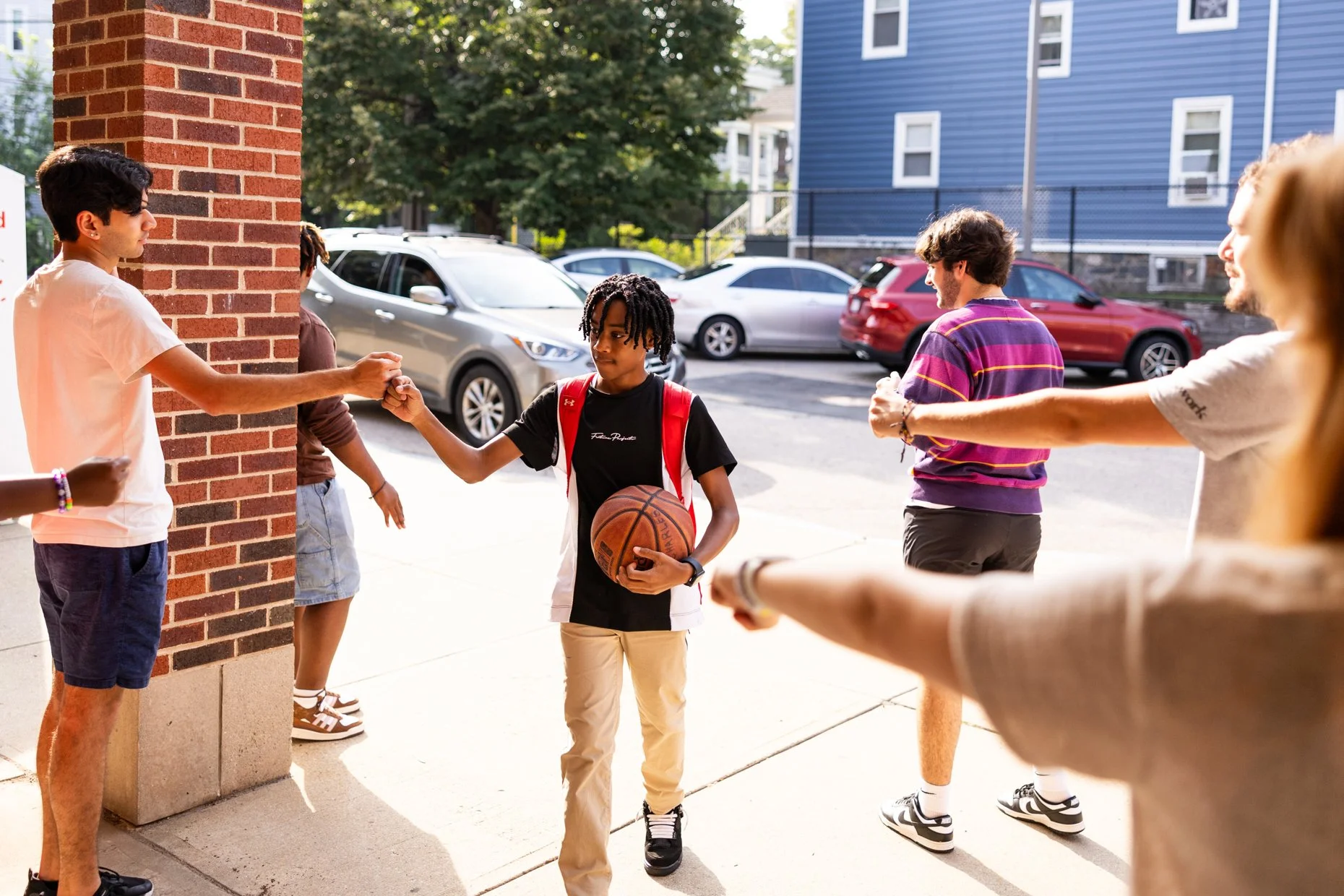 Teen boy with a basketball, giving a fist bump to a young man near a brick wall, with others around in a school or community setting outside, parked cars in background.