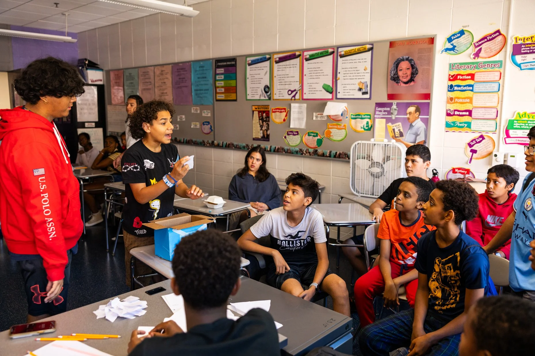 A classroom scene with a teacher and students. The teacher, wearing a red hoodie, is standing on the left side, while a girl is standing and speaking to the group. Several students are sitting at desks, listening and looking at the speaker. The class