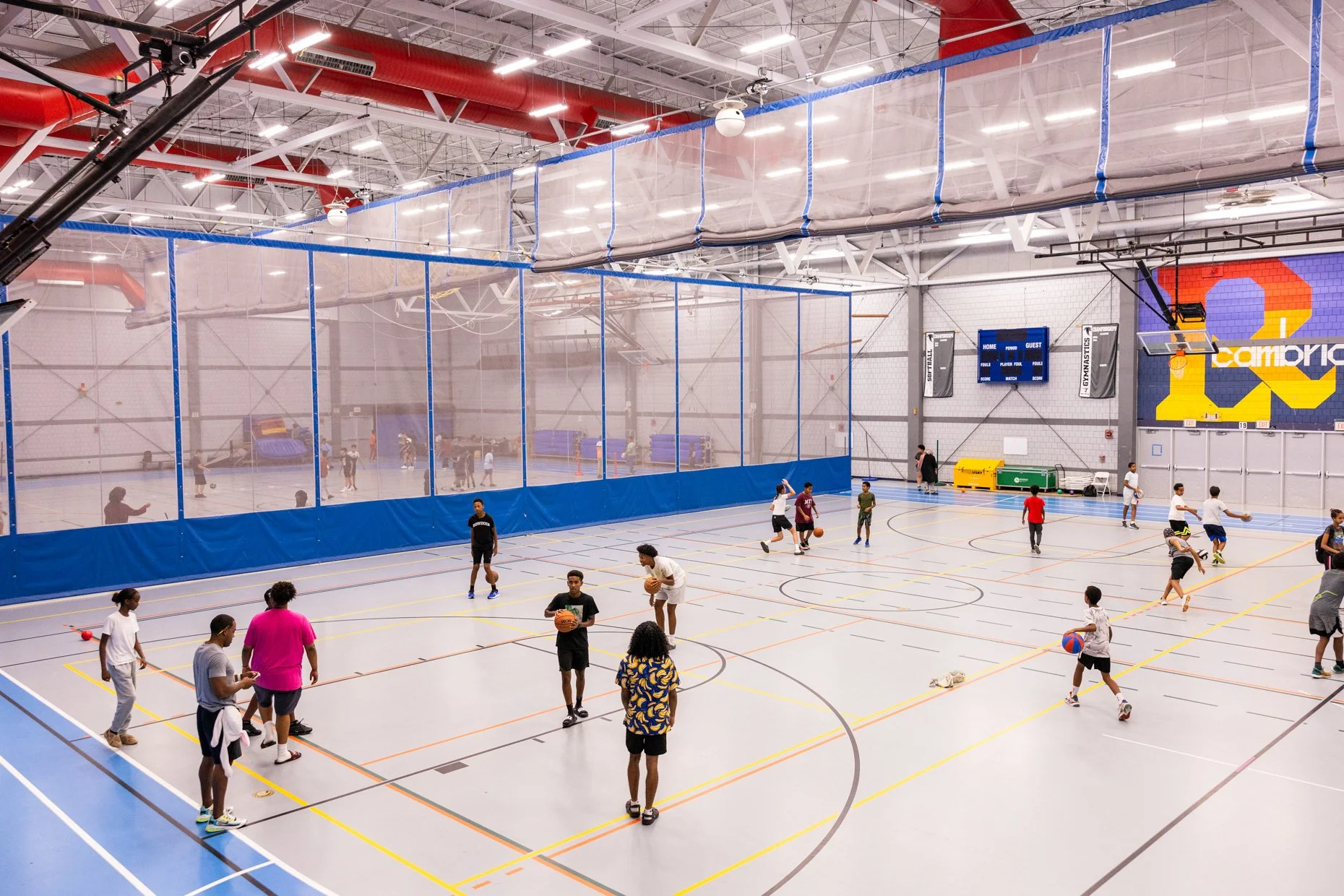 An indoor gymnasium filled with children and teenagers playing basketball and other sports, with a large colorful scoreboard and a court divided by a high net.