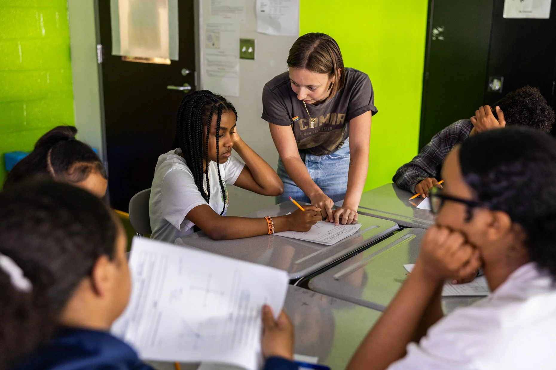 A teacher assists a student during a classroom activity, with students seated around a table, some reading papers, in a brightly colored classroom with green walls.