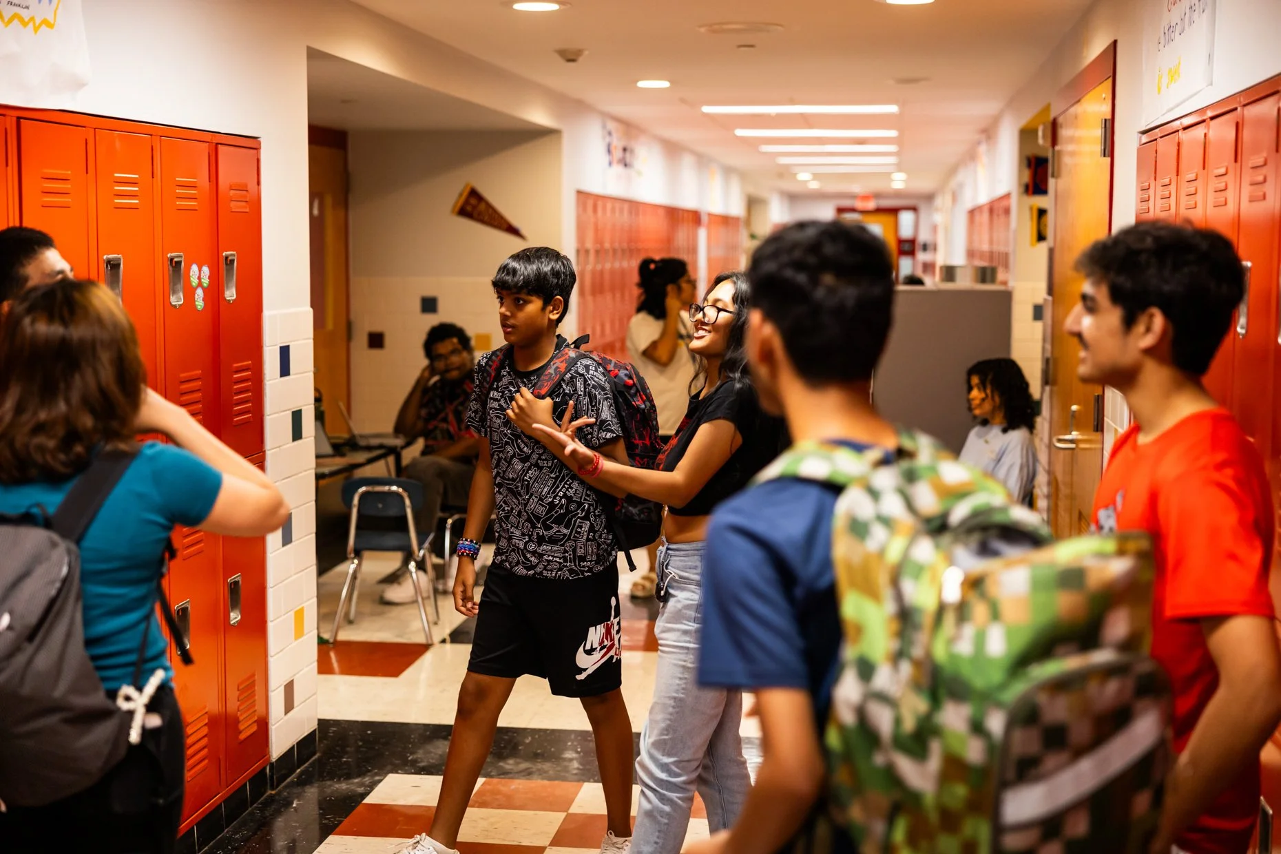 Students in a school hallway near lockers, with some talking and using a cellphone, colorful lockers, and students with backpacks.