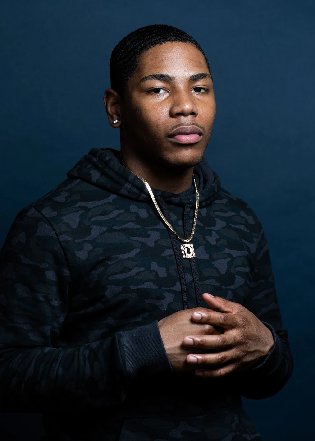 Portrait of a young man with a serious expression, wearing a black camo hoodie, gold chain with a 'D' pendant, diamond earrings, and his hands crossed over his chest, against a dark blue background.