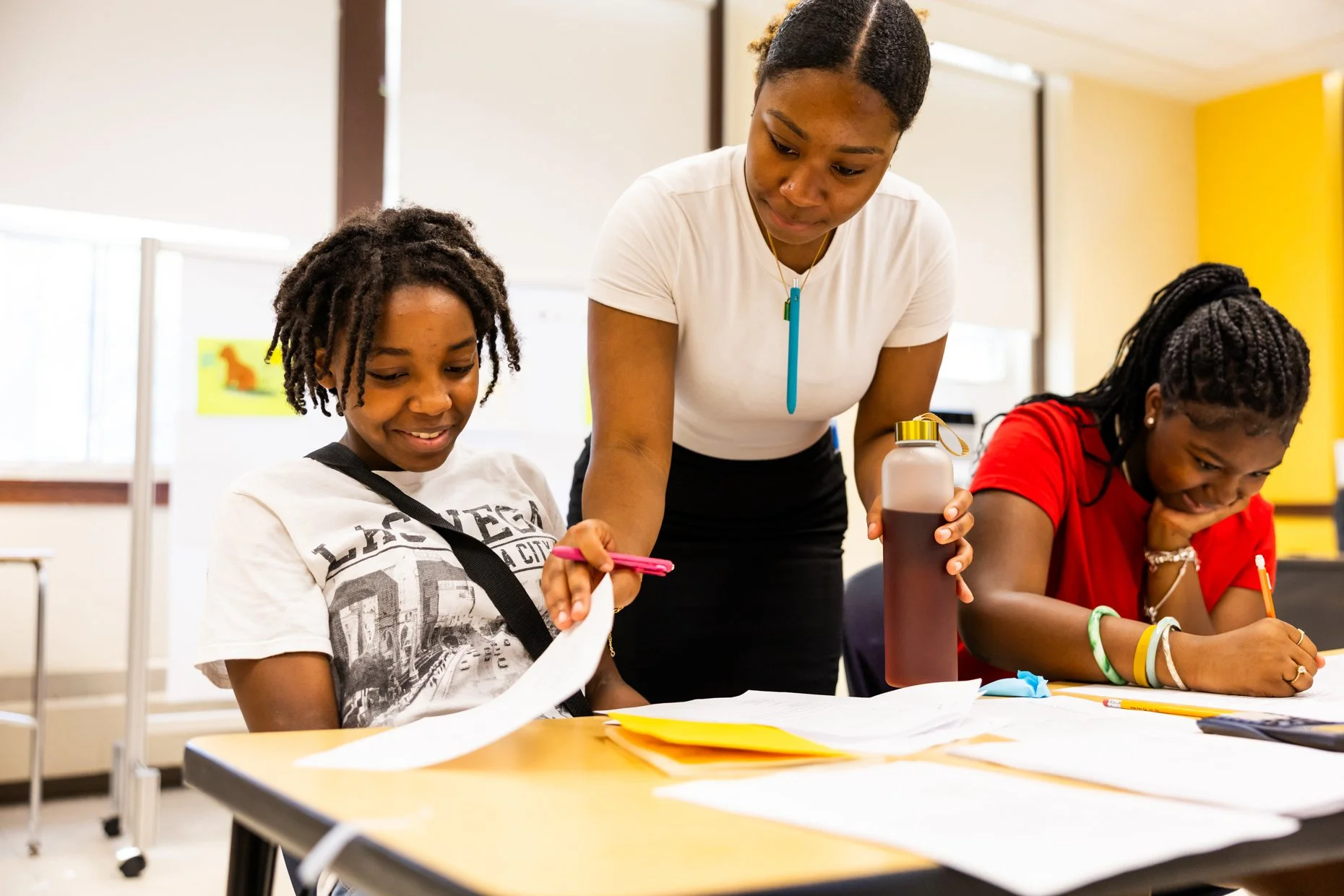 A teacher assists two students in a classroom, one holding a pink marker and the other writing on paper, with papers, colorful pens, and a bottle of juice on the table.
