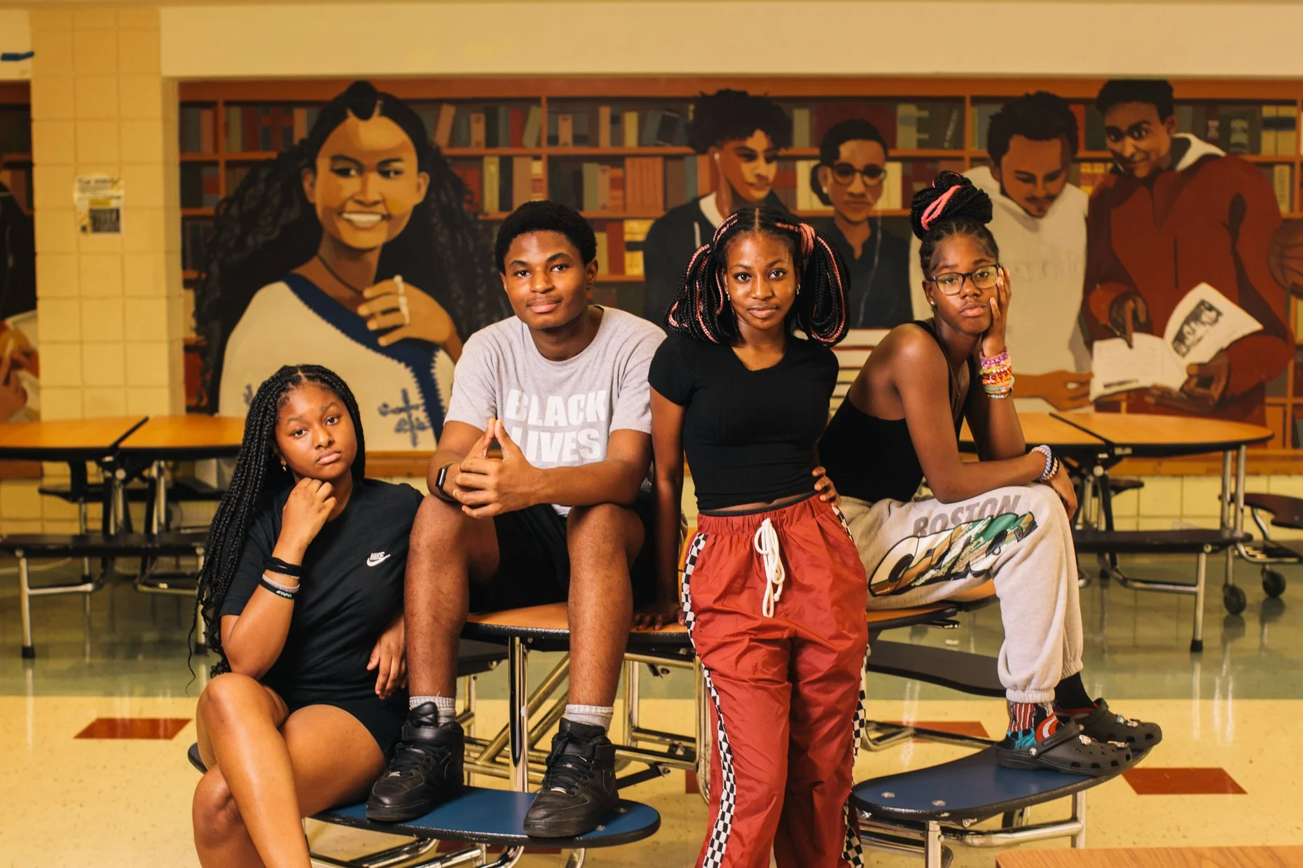 A group of five diverse young people posing in a school cafeteria, with a large mural of children painted on the wall behind them.