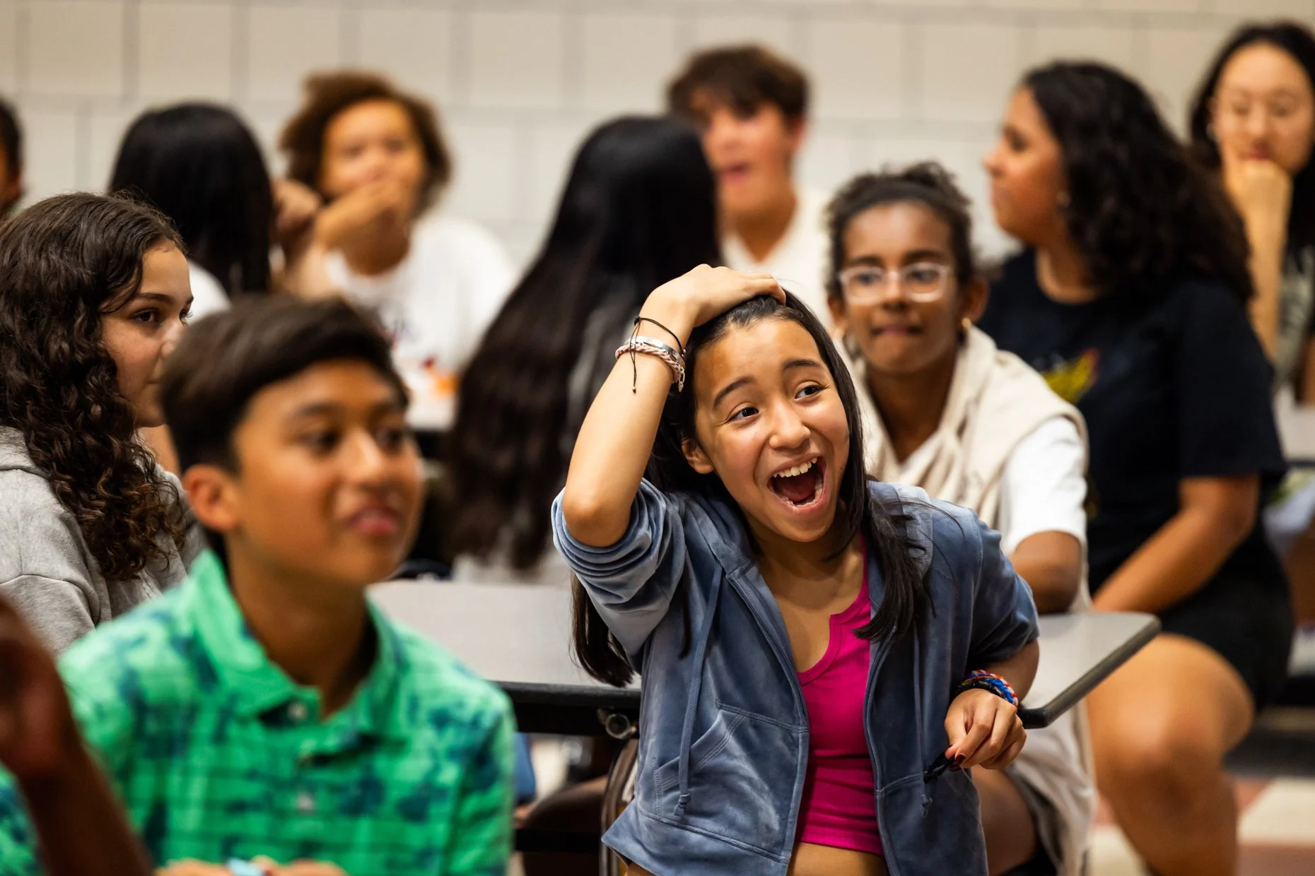 A group of children sitting in a classroom, with one girl in the center laughing and touching her hair, surrounded by classmates who are engaging and smiling.