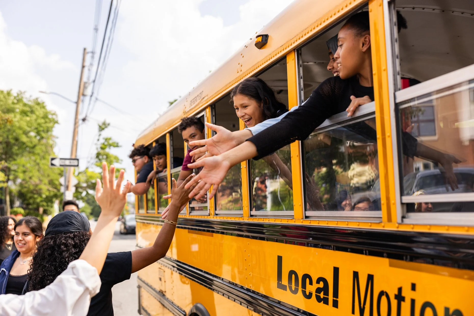 Young people on a yellow school bus reaching out to a crowd below, smiling and waving during daytime, with trees and power lines in the background.