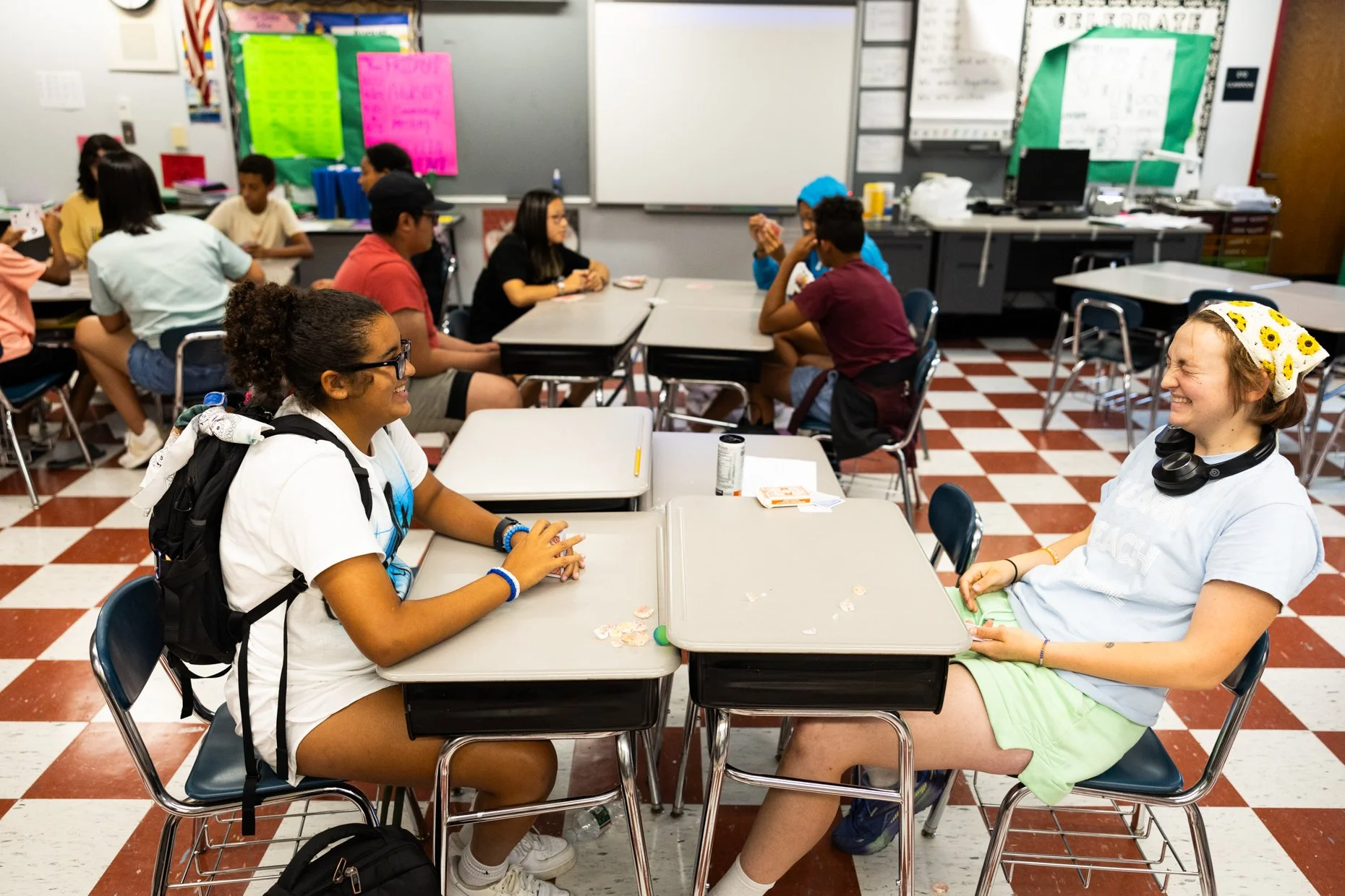 Students sitting and chatting at desks in a classroom, some laughing and smiling, with classroom decorations and supplies in the background.
