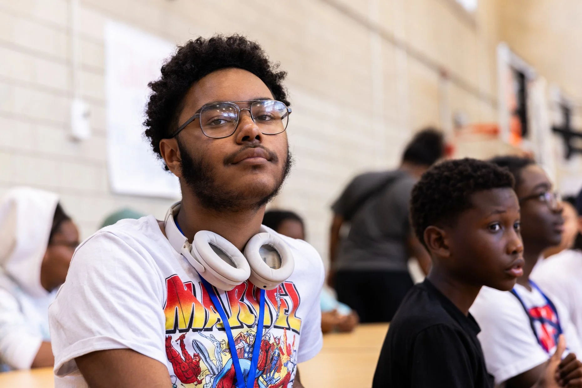 Young man with curly hair, glasses, and headphones around his neck sitting in a classroom with other students.