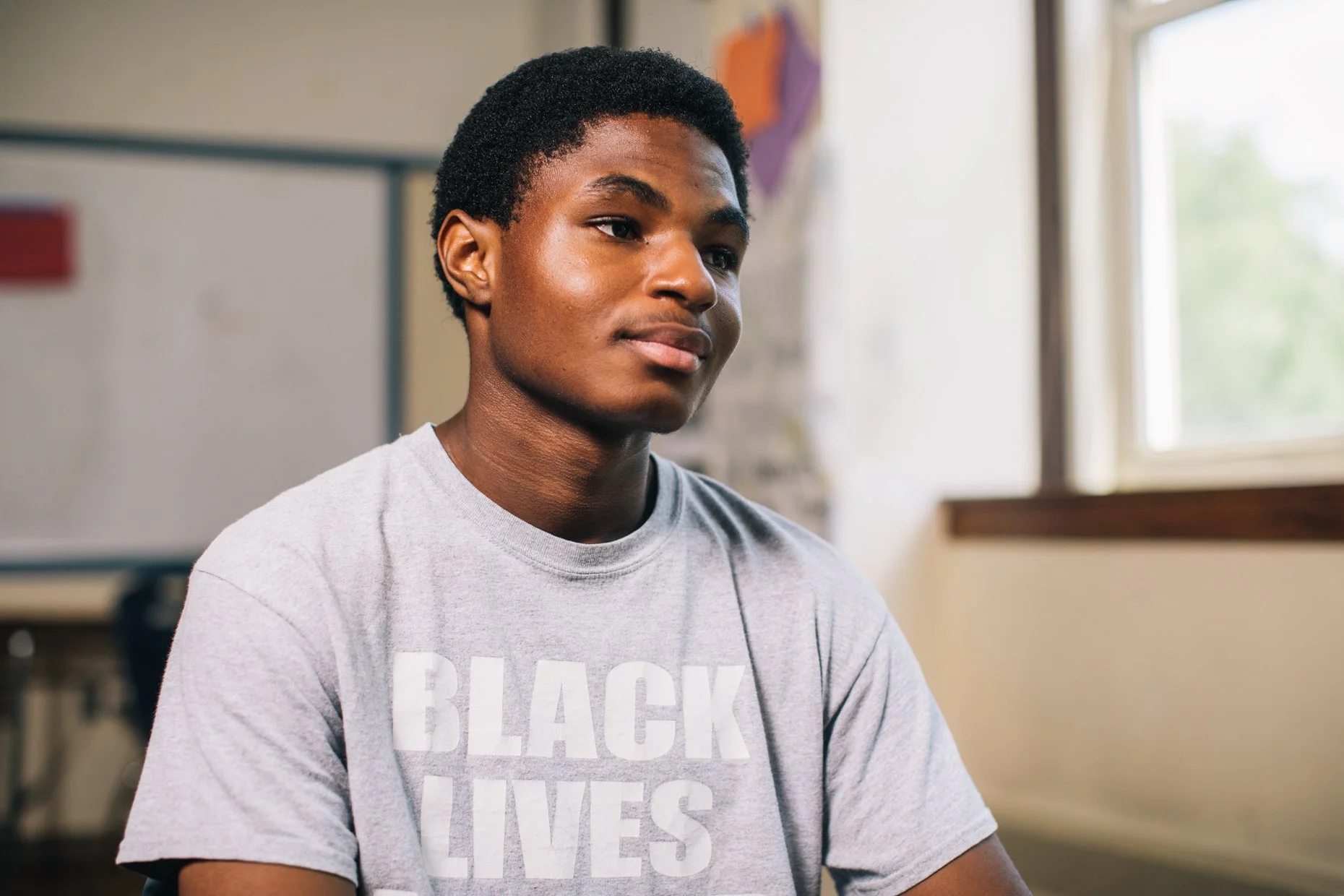 A young Black man with short curly hair, wearing a gray t-shirt with white text, sitting in a classroom or indoor space near a window, looking slightly to the right.