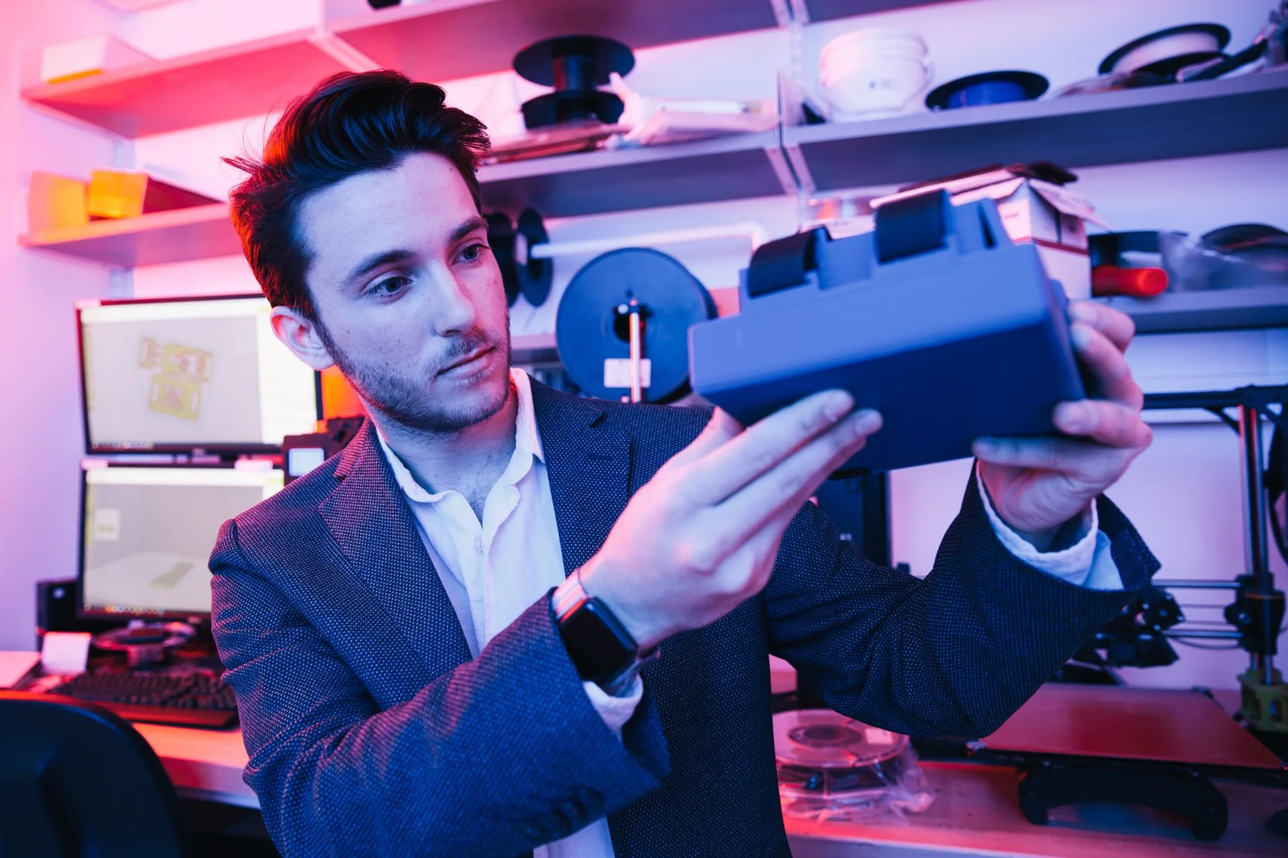A young man with dark hair and a beard, wearing a dark blazer and a white shirt, is inspecting a small electronic device or remote control in a high-tech workspace. The background includes computer monitors, shelving with electronic equipment, and co