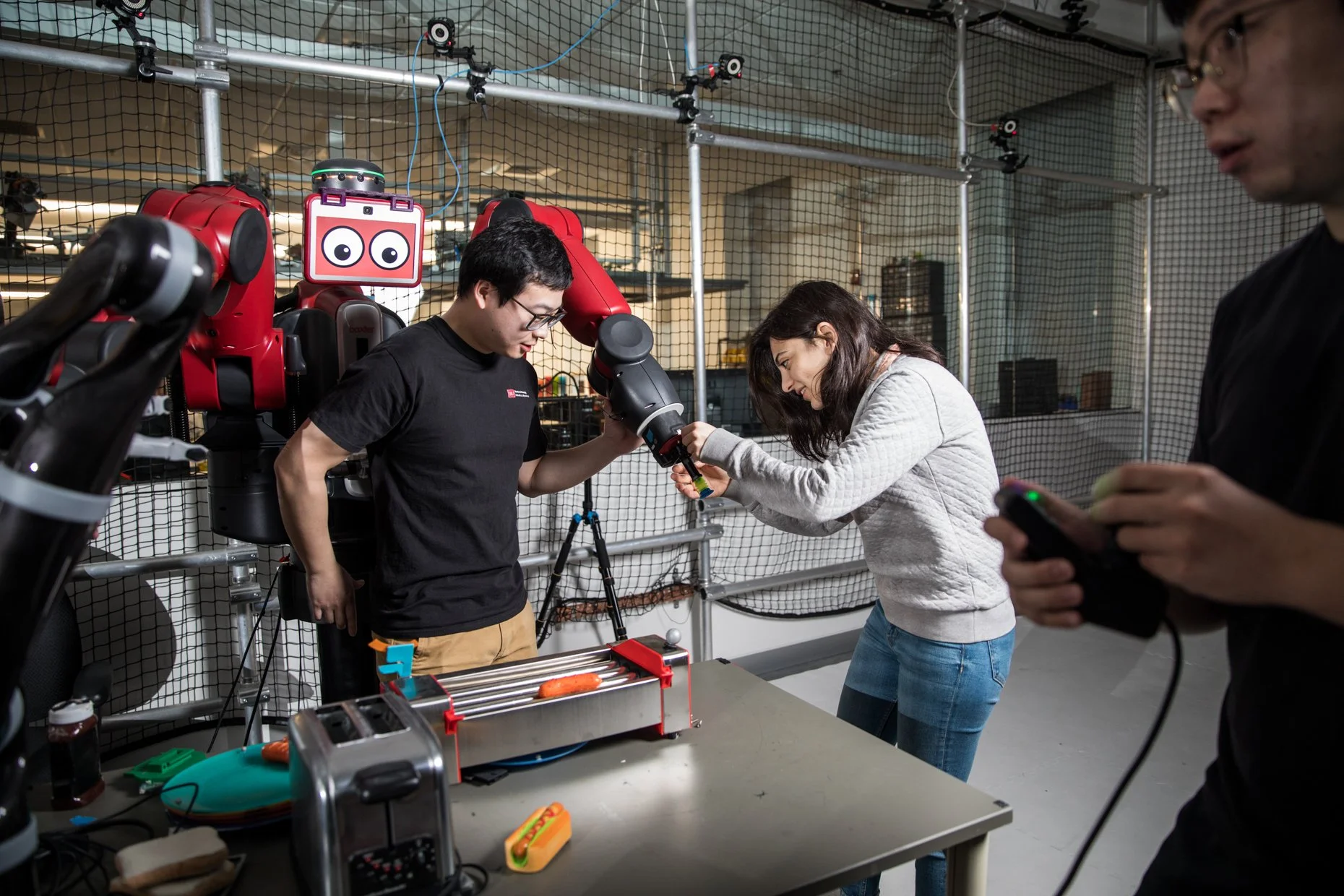 People working on a robotics project in a workshop. One person is holding a robotic arm, another is adjusting a component, and a third is holding a remote control. The workspace includes various tools and equipment, with a robot and sensors visible i