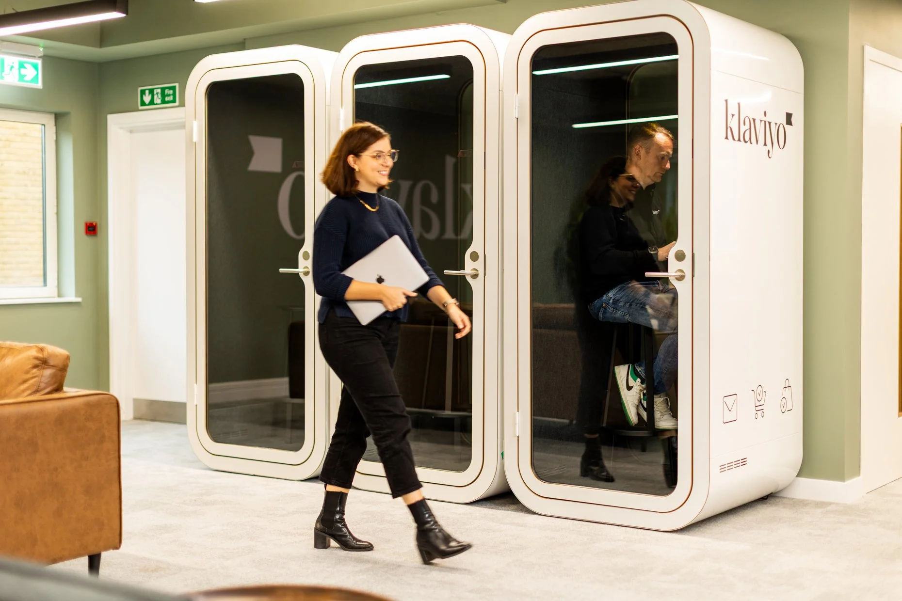A woman walking past a phone booth-like structure labeled 'klaviyo' with a person sitting inside using a laptop.