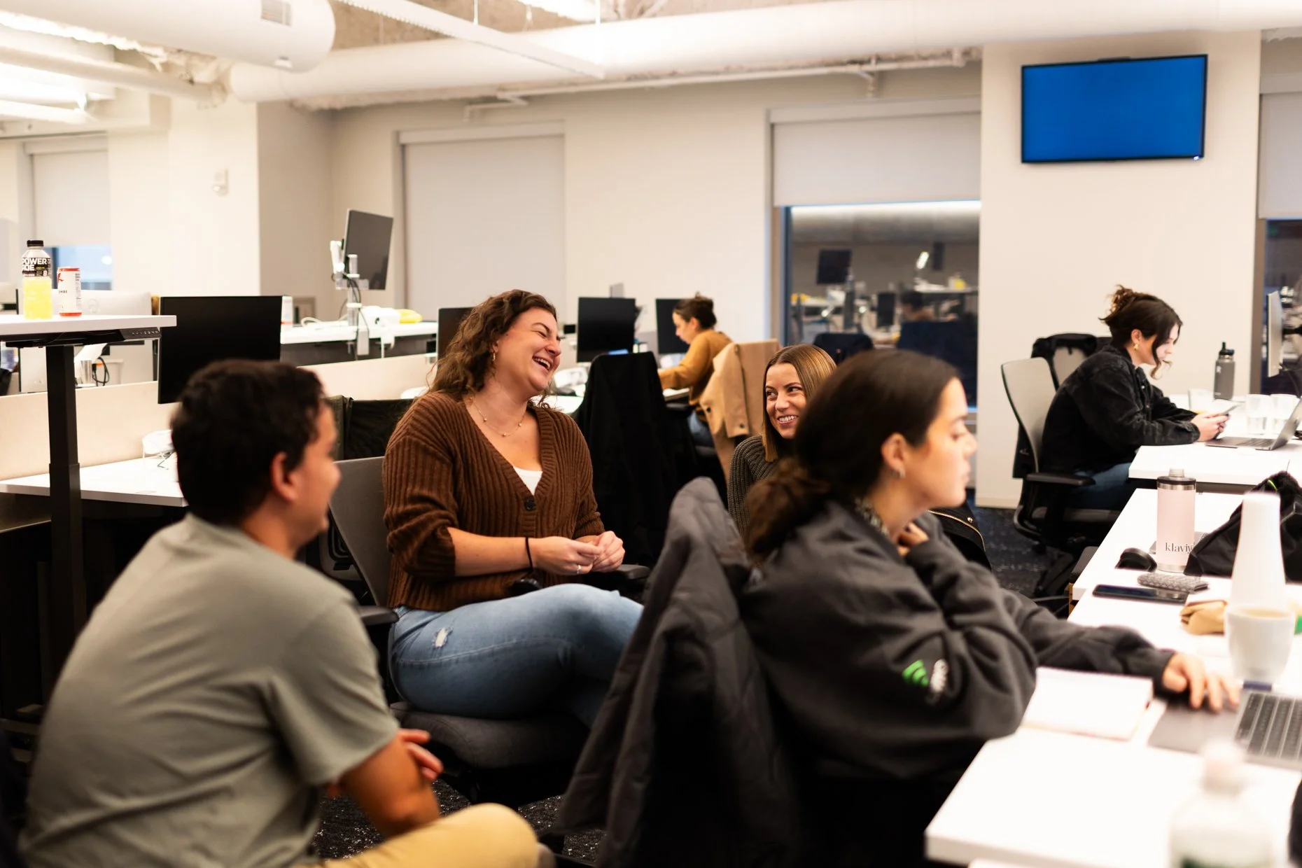 Group of people in a modern office space engaging in conversation and working on their laptops.