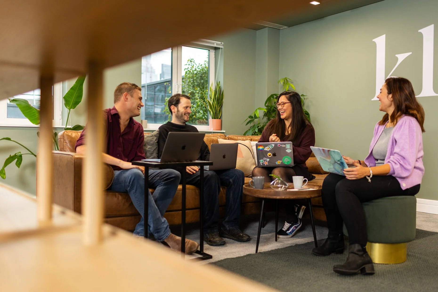 Four people sitting together in a cozy room, engaged in conversation and smiling. Three women and one man, with laptops and tablets, sitting on a couch and chair around a small table with coffee cups, surrounded by green plants and sunlight coming th