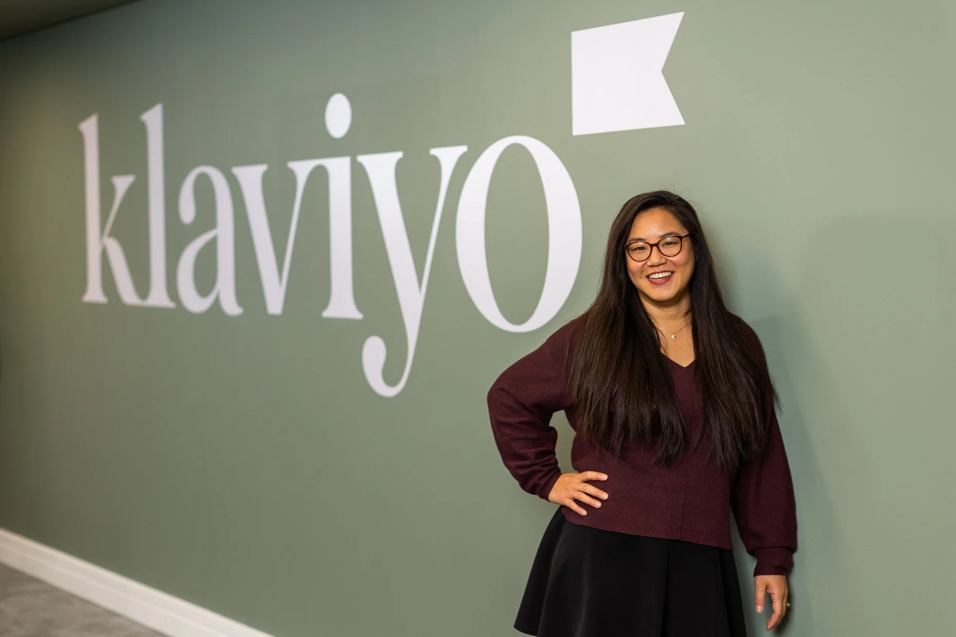 A woman with long dark hair, glasses, wearing a maroon top and black skirt, standing in front of a green wall with the word 'klavyo' and an arrow logo, smiling with her hand on her hip.