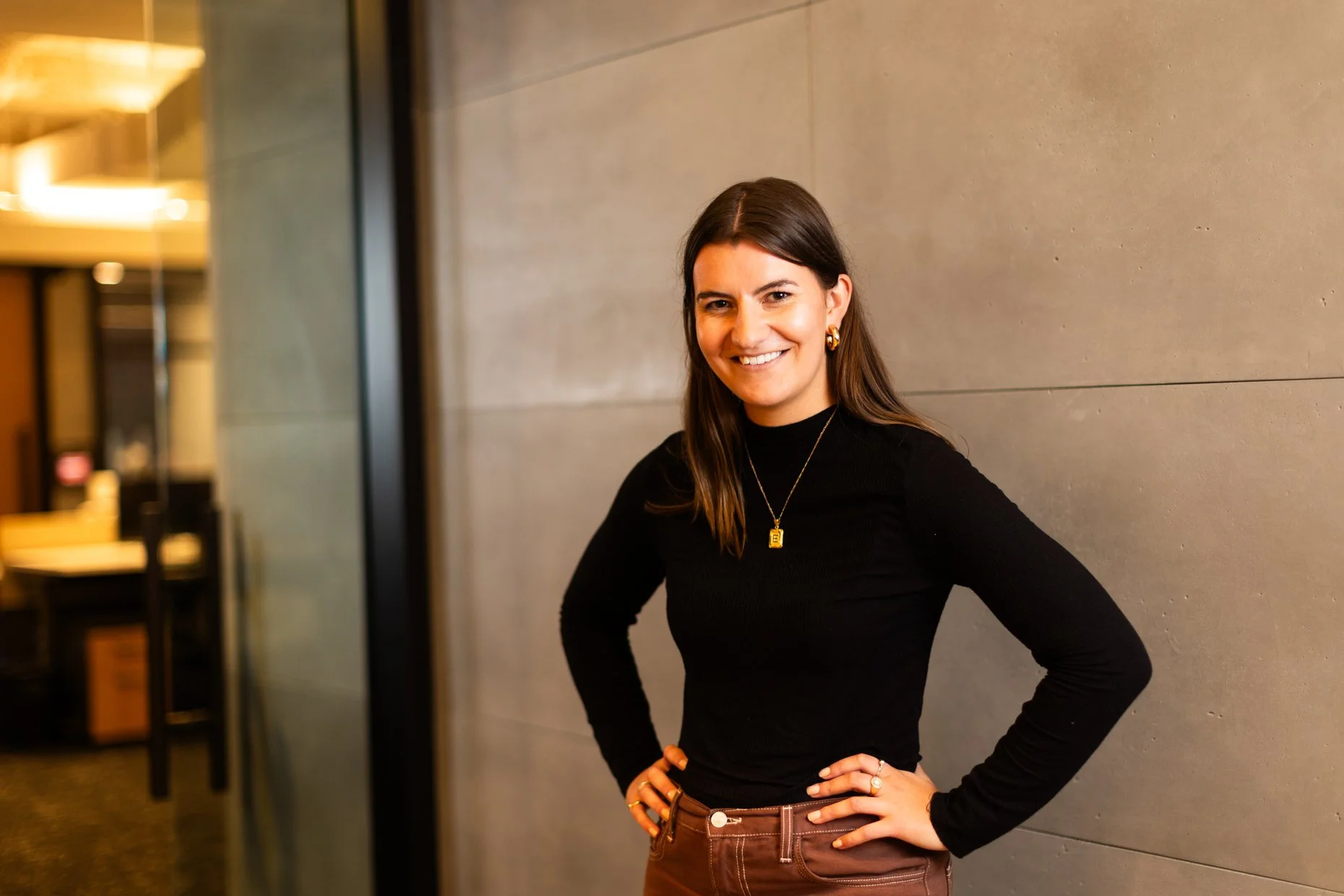 A woman with long dark hair, wearing a black top and brown pants, posing with her hands on her hips and smiling in an indoor setting.