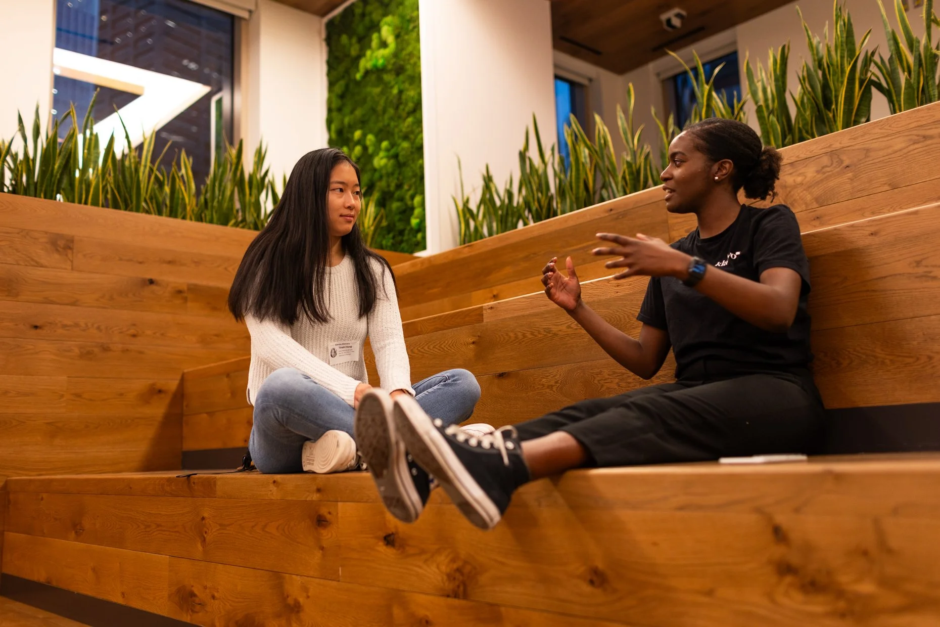 Two young women are sitting on wooden steps indoors, engaged in conversation. One woman with long dark hair is listening attentively while the other woman with short curly hair is speaking.