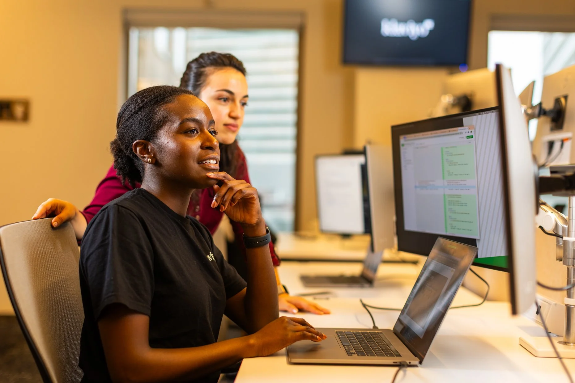 Two women working together at a computer in an office. One woman is seated, looking at the monitor with a thoughtful expression, while the other woman stands behind her, observing. Multiple computer screens and a TV monitor are visible in the backgro