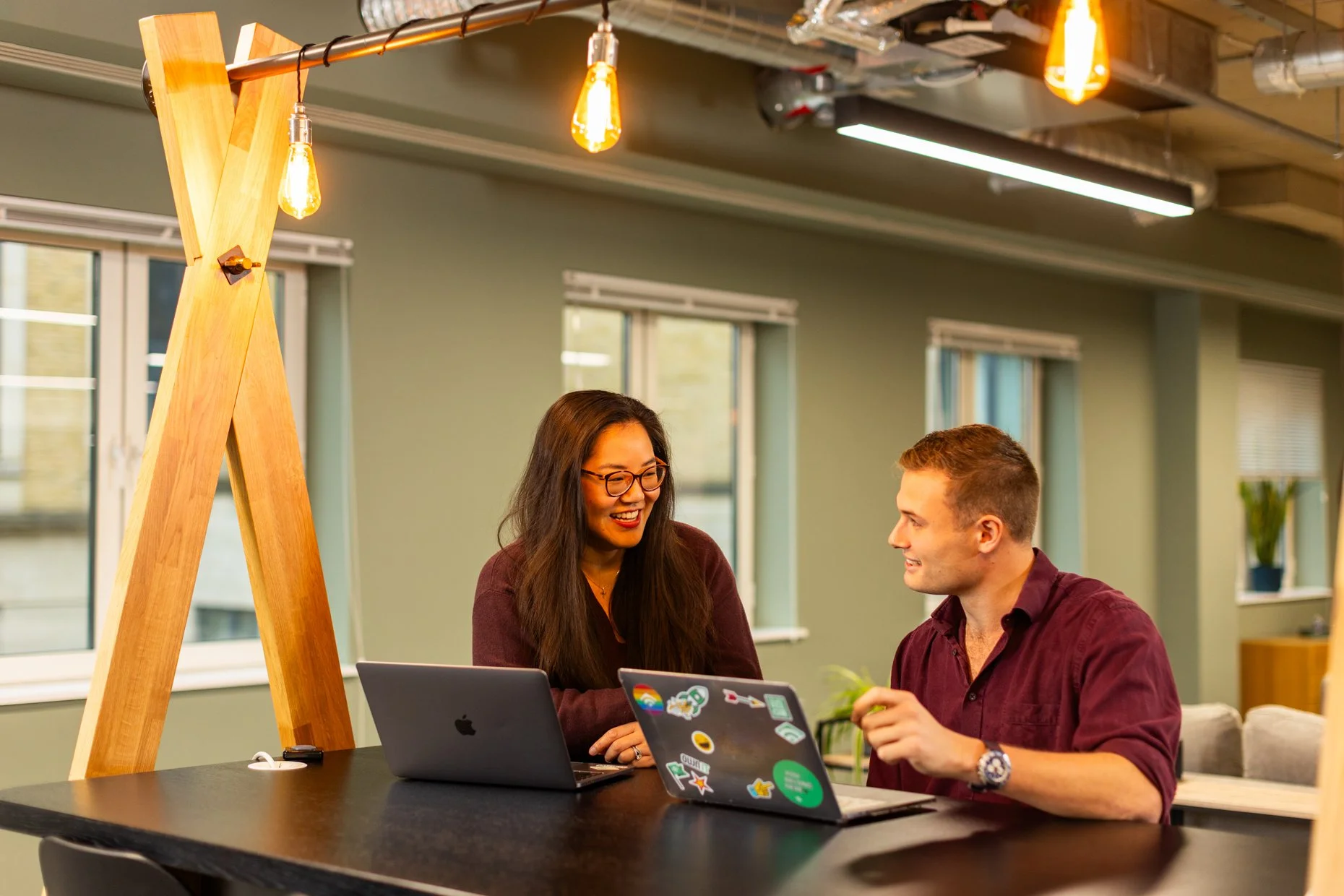 Two people, a woman and a man, are sitting at a black table with laptops in a modern, brightly lit office space. The woman has long dark hair, glasses, and is smiling, while the man has short hair and is smiling back. The woman’s laptop is open in fr