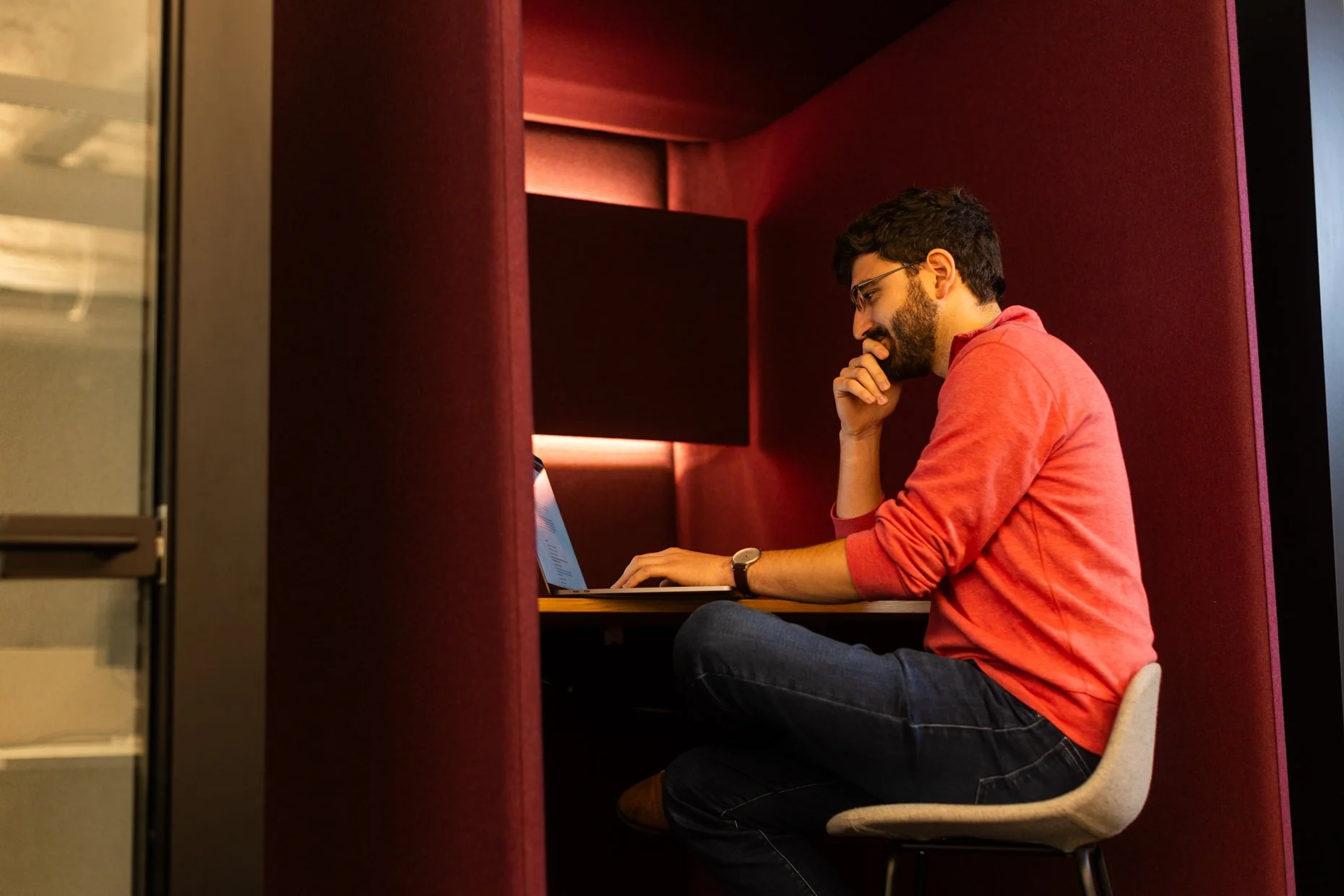 A man with glasses and a beard, sitting in a booth with red walls, working on a laptop, wearing a red shirt with rolled sleeves and a wristwatch.