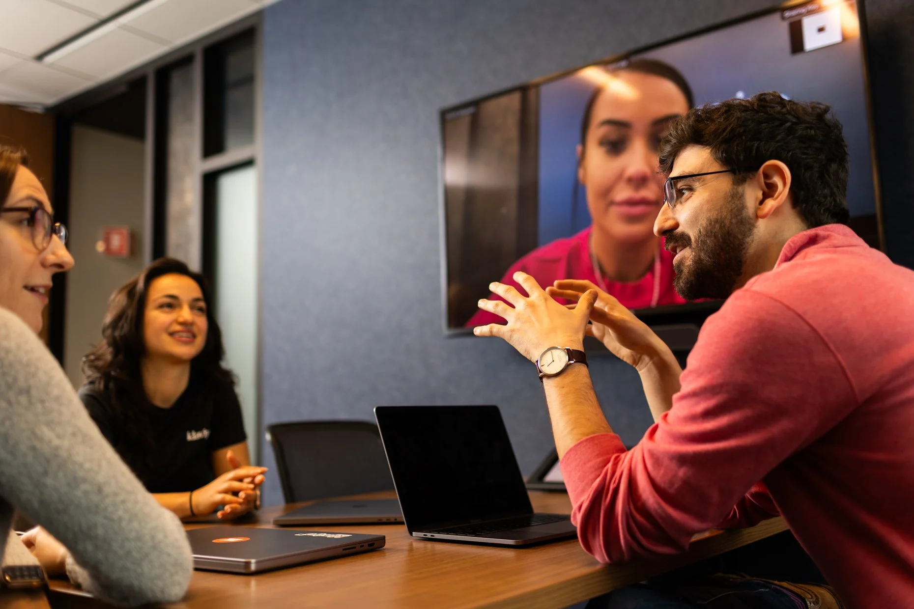 A group of four people having a discussion in a meeting room, with one person on a video call displayed on the screen.