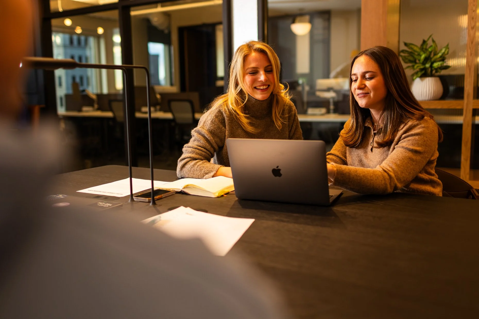 Two women sitting at a table working on a laptop in a modern office setting, with notebooks, papers, and pens on the table, and large windows in the background.