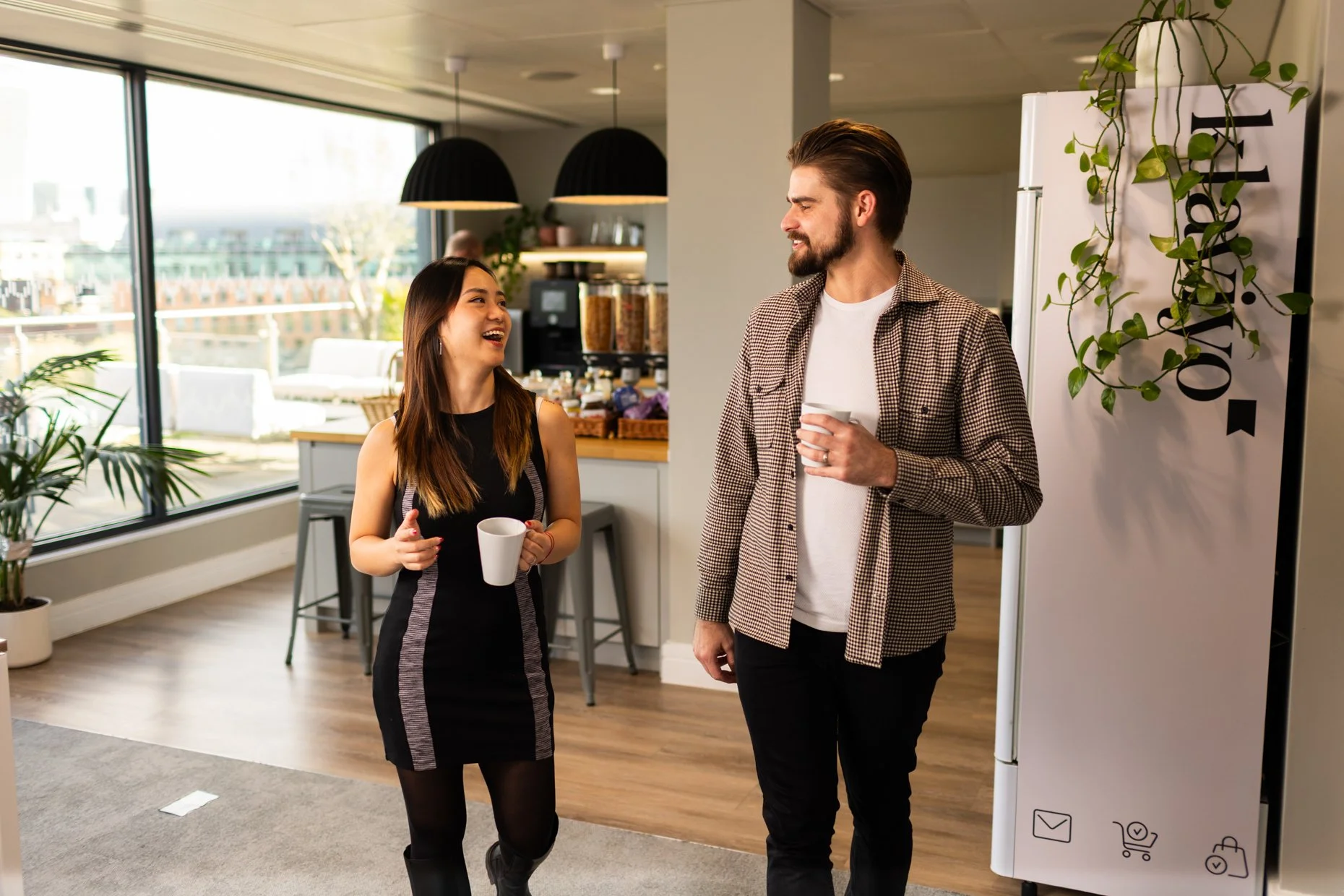 A man and woman are smiling and talking while holding coffee mugs in an office space with large windows, indoor plants, and modern decor.