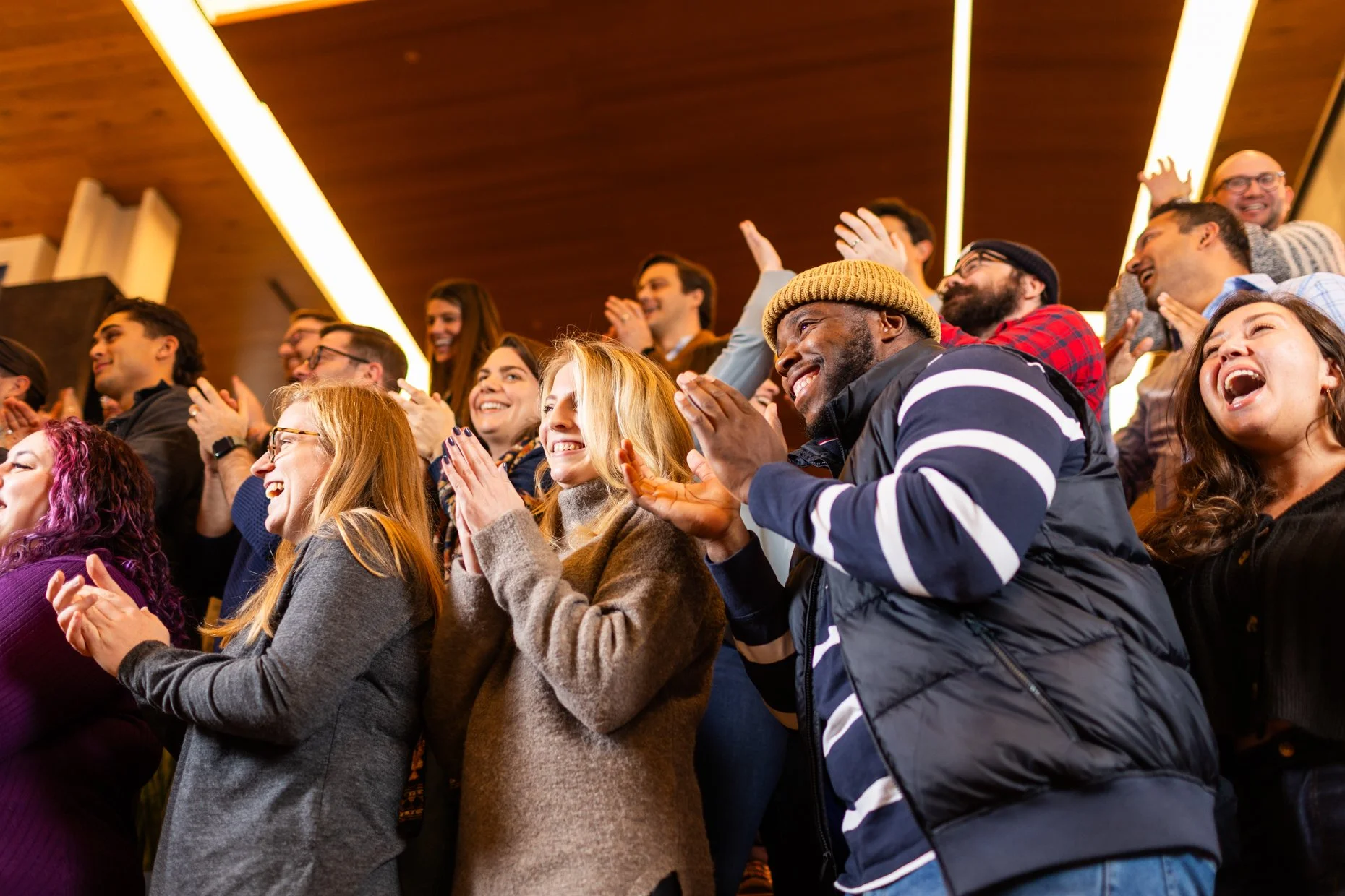 A group of diverse people in an audience, clapping, smiling, and laughing at an event, under warm lighting with wooden accents on the ceiling.