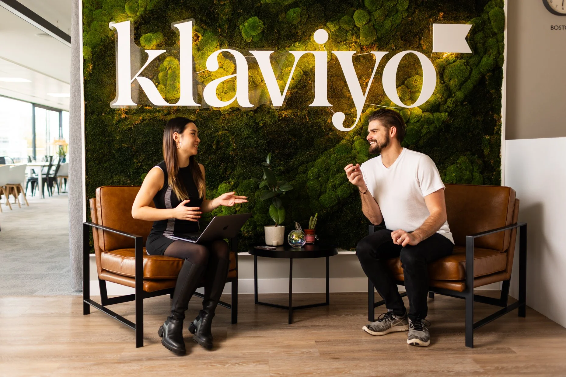 Two people sitting and talking in a modern office with a green moss wall and a large sign that reads 'klaviyo' in the background.
