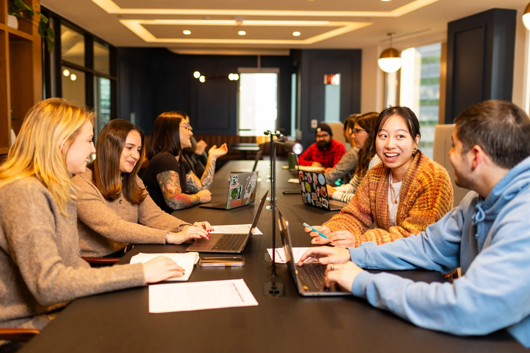 Group of young adults sitting around a conference table in a modern office, engaged in conversation and using laptops and smartphones, with a bright, well-lit room in the background.
